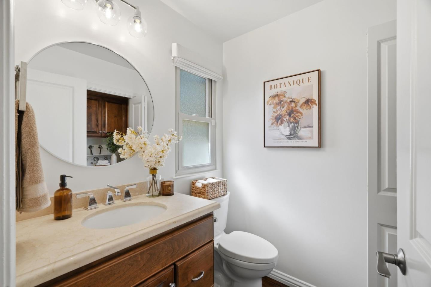 Bathroom with dark wood vanity, oval mirror, frosted window, and framed floral print.