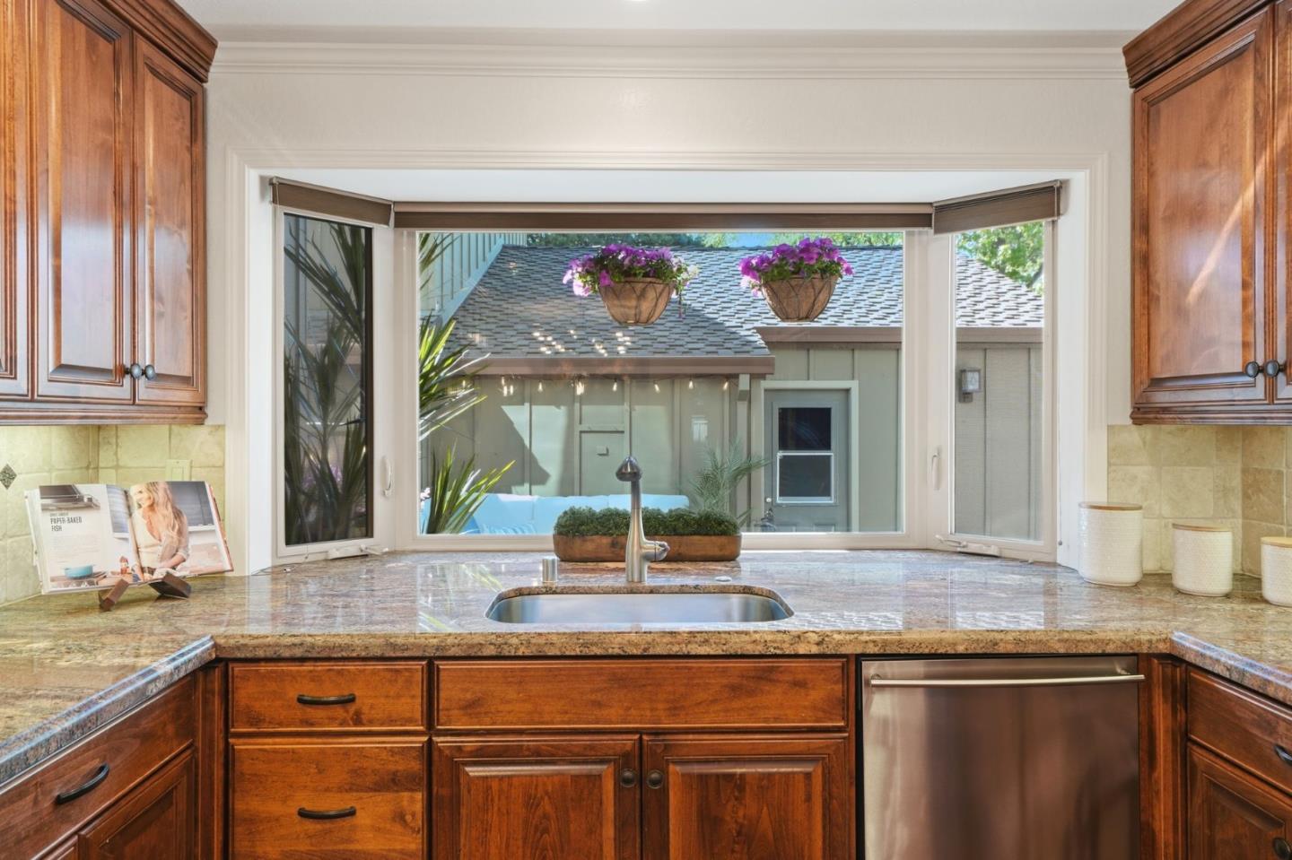 Kitchen sink beneath a wide courtyard-facing window, framed by wood cabinets and granite countertops.