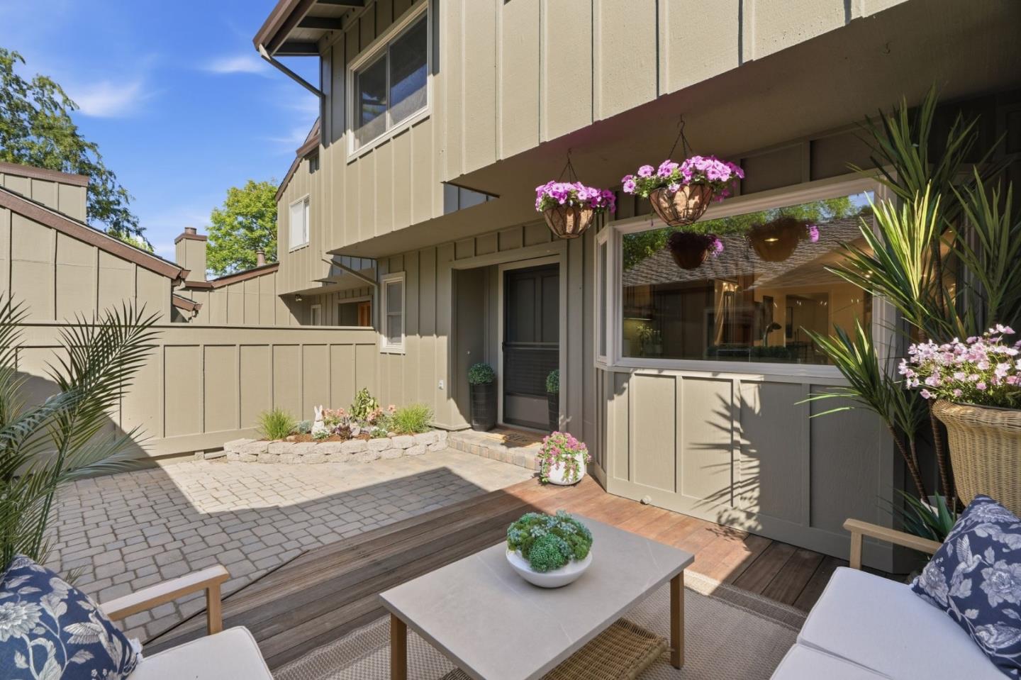 Patio with cushioned seating, flowering planters, paved courtyard, and a large window.