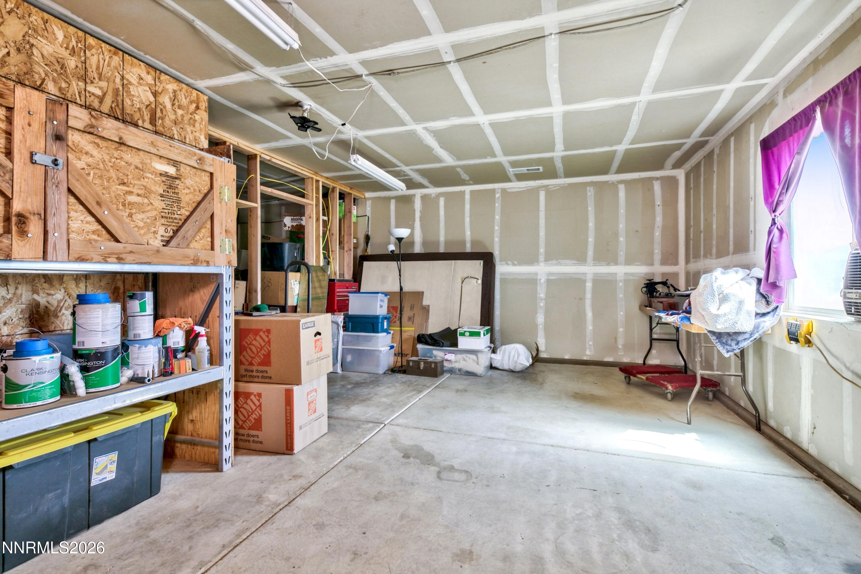 Garage with unfinished drywall, storage shelves, paint cans, and boxes beside a curtained window.