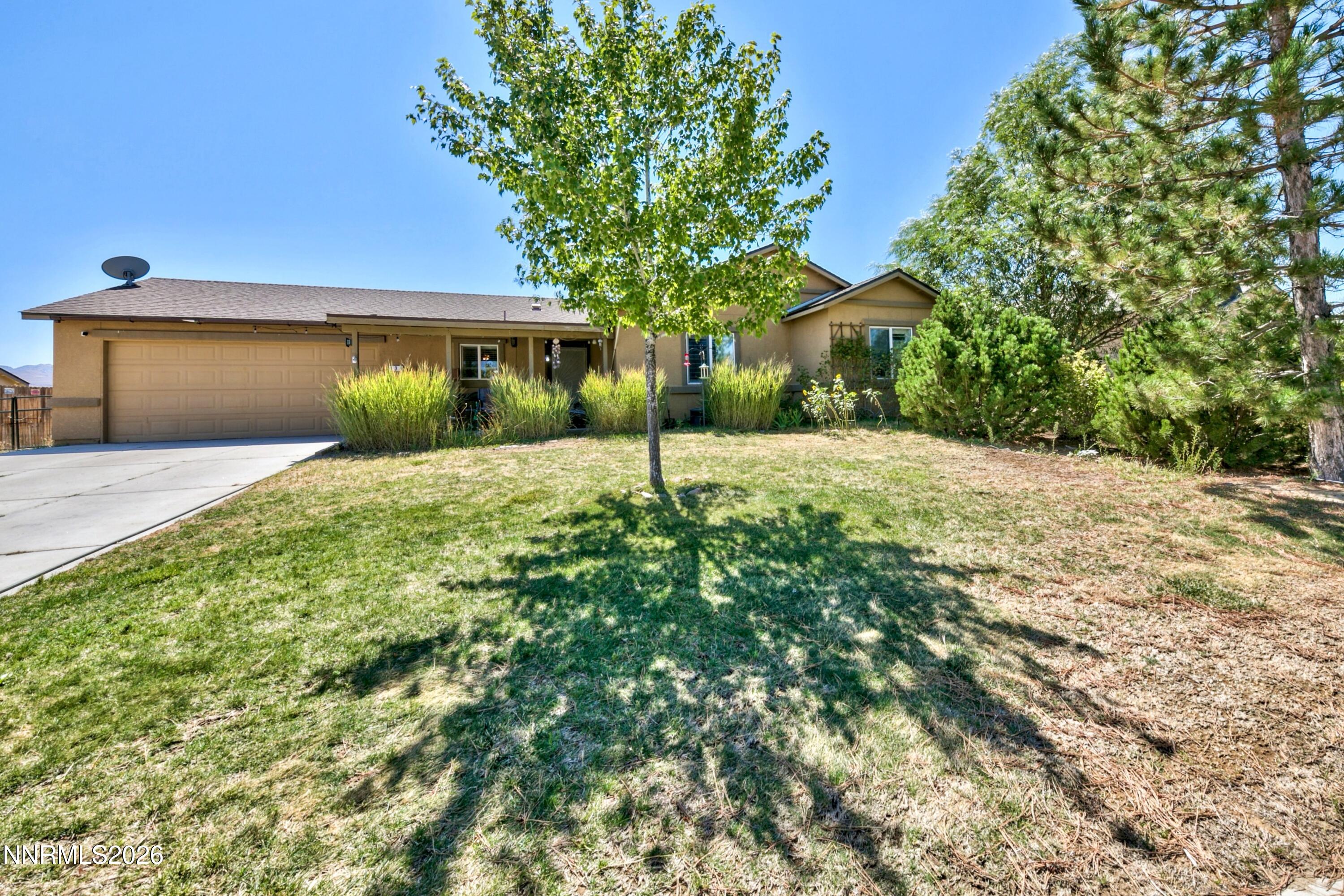 Single-story tan home with a two-car garage, front lawn, ornamental grasses, and mature evergreens.