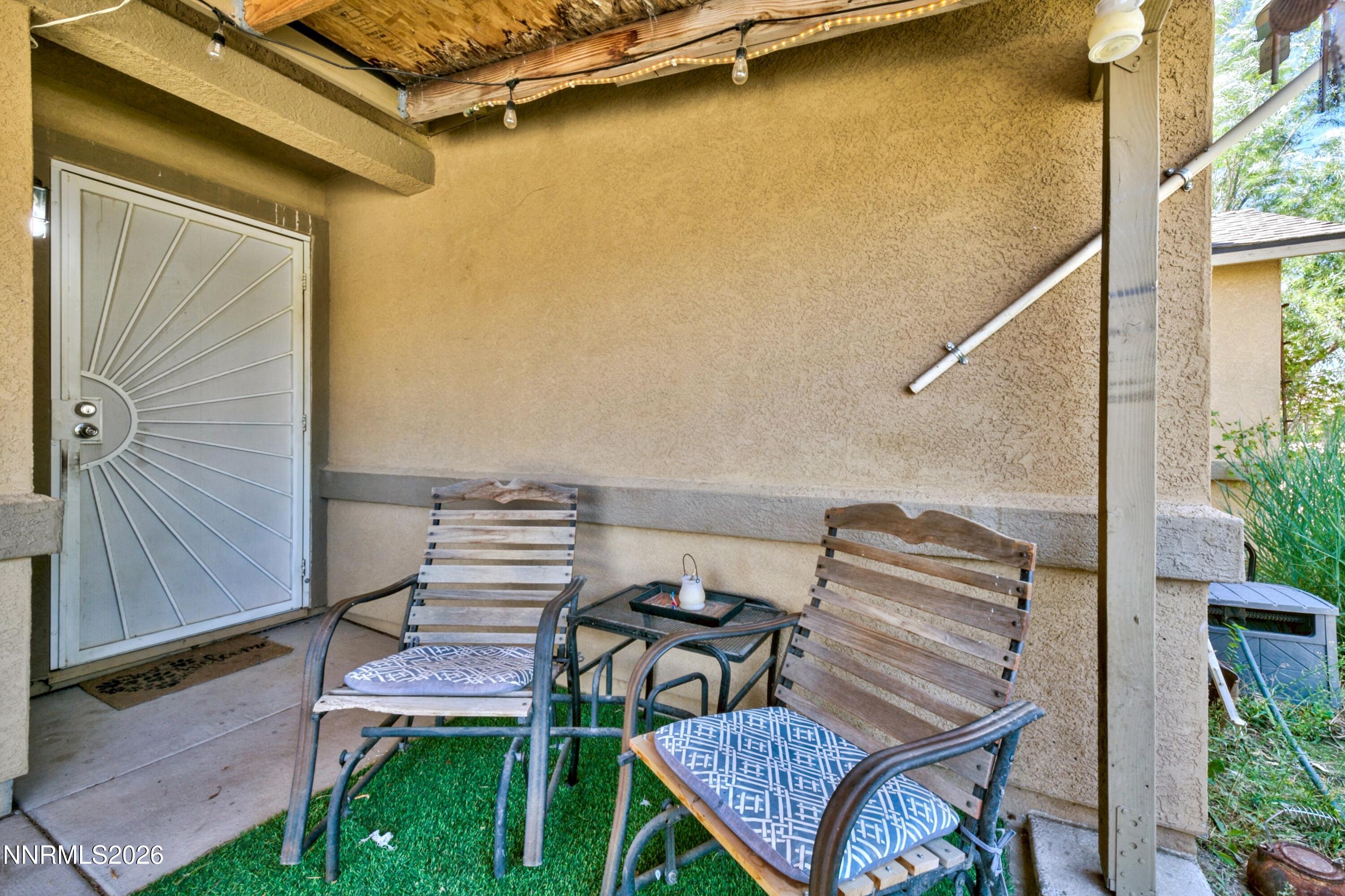 Covered porch with two wooden chairs, patterned cushions, a small table, and string lights.
