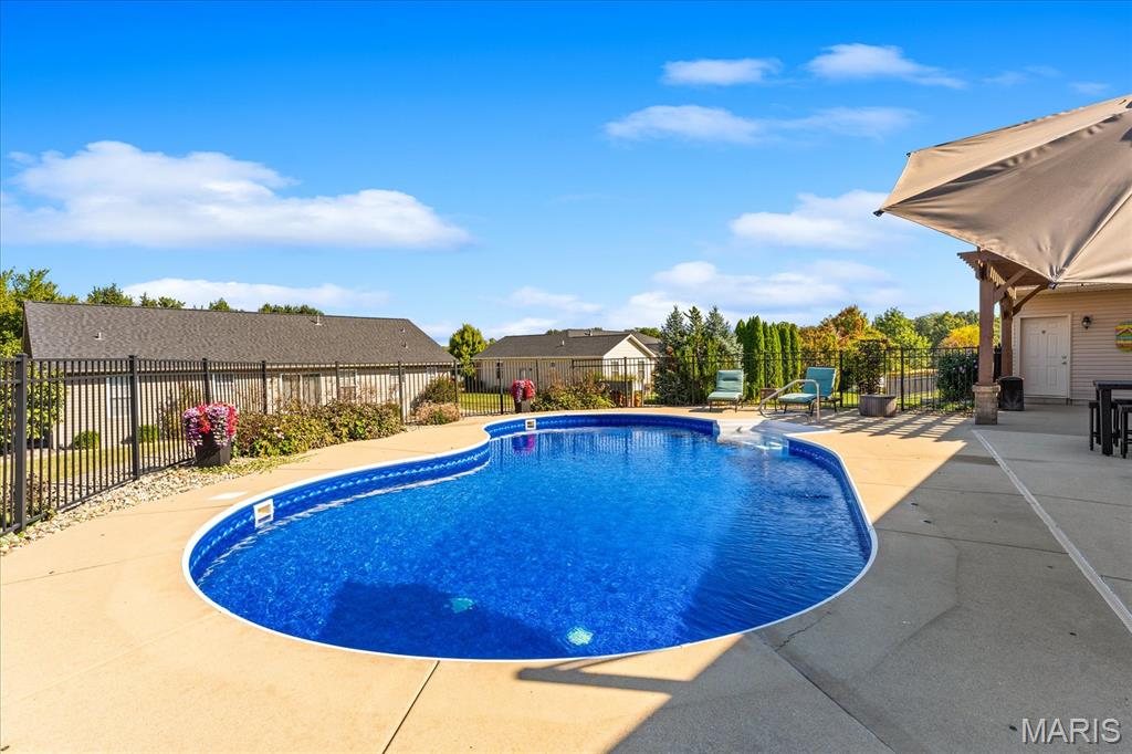 Curved blue swimming pool bordered by a concrete patio, black fencing, and a covered seating area.