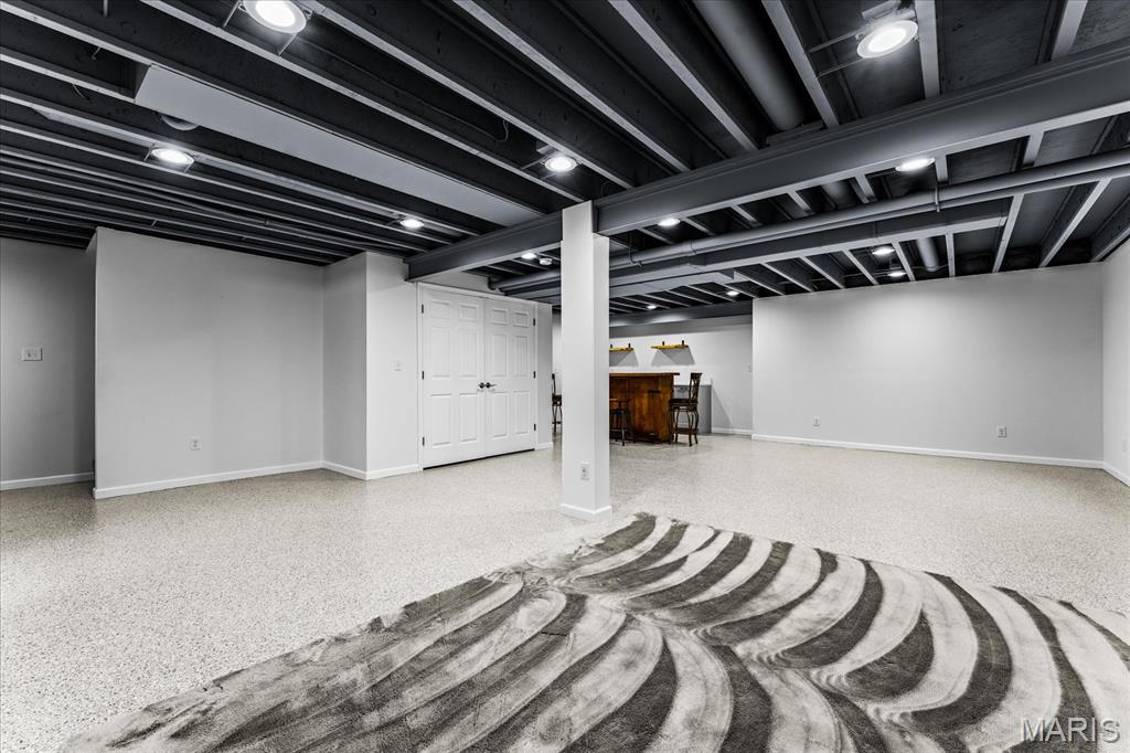 Finished basement with exposed ceiling beams, a small bar, and a large patterned rug.