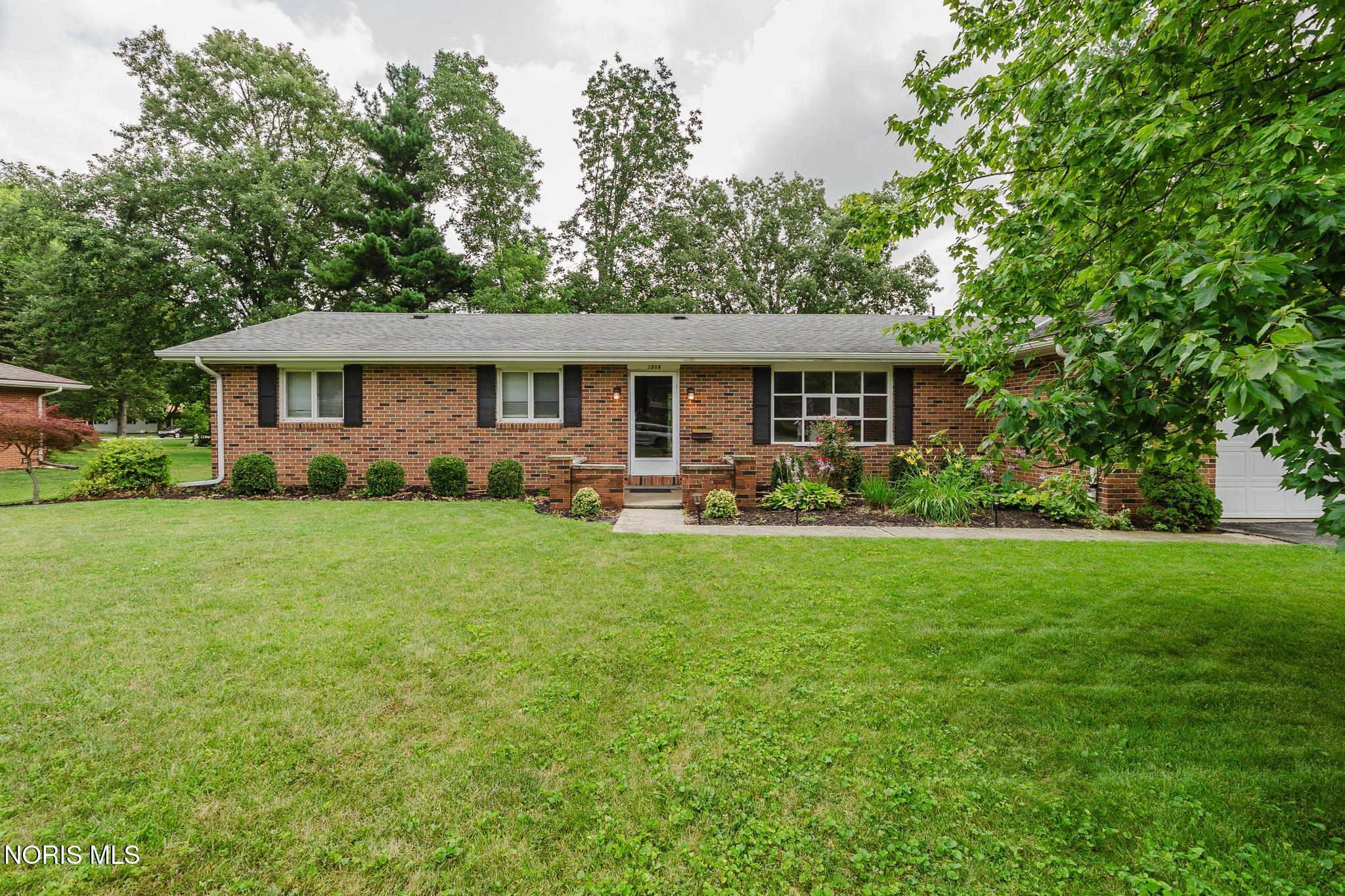 Brick ranch house with a broad lawn, mature trees, and landscaped flower beds.