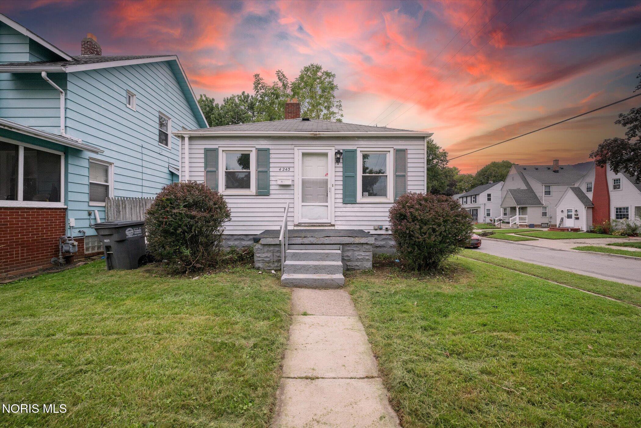 White-sided house with green shutters, front steps, and a walkway under a vivid sunset sky.