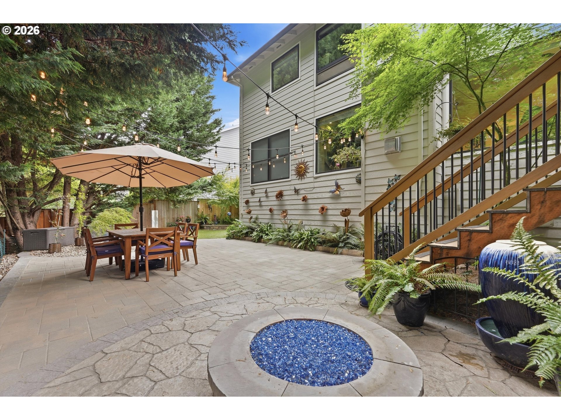 Backyard patio with umbrella-covered dining set, string lights, garden steps, and a circular blue fire feature.