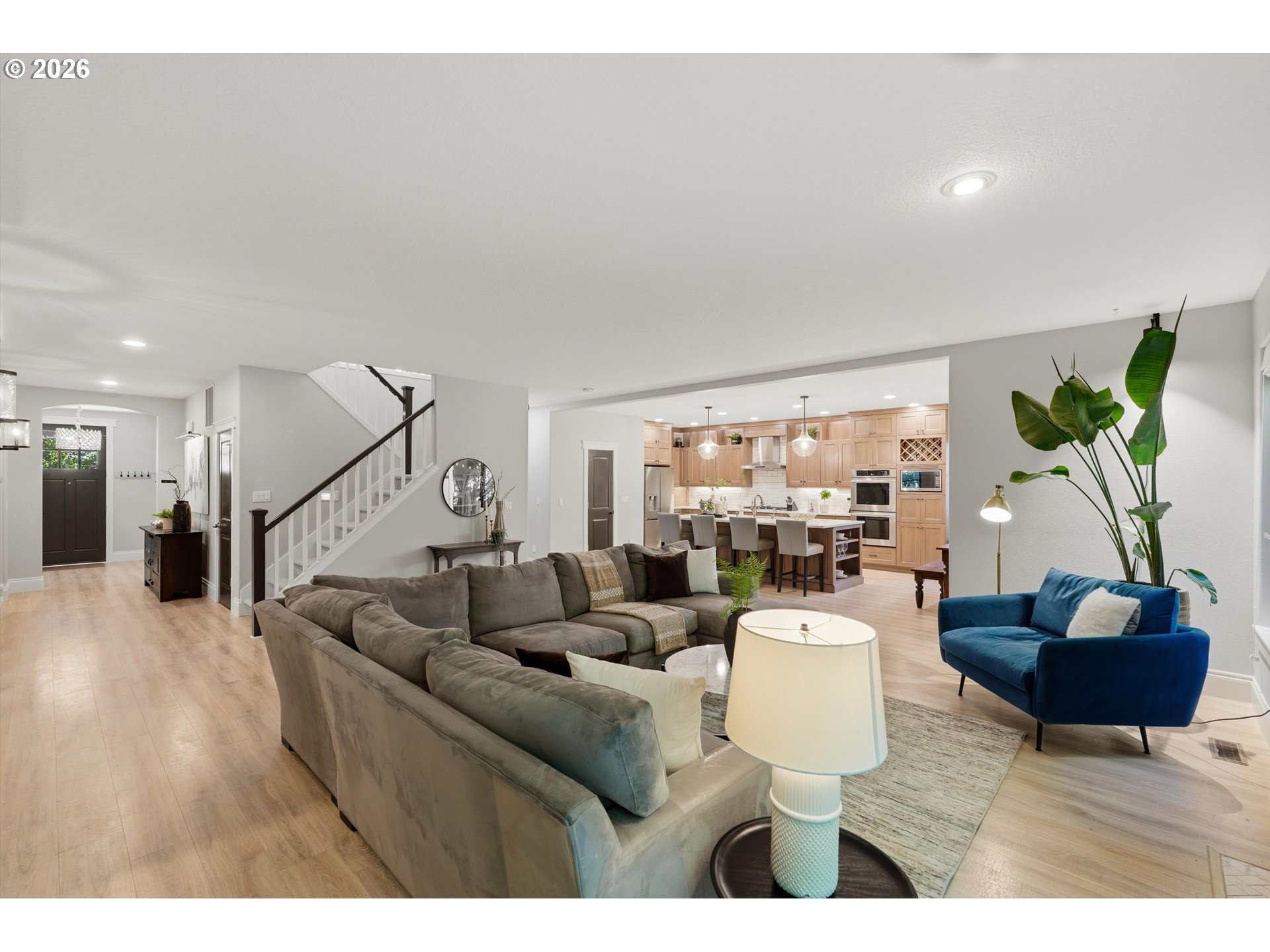 Open-plan living room with gray sectional, blue armchair, staircase, and light wood kitchen.