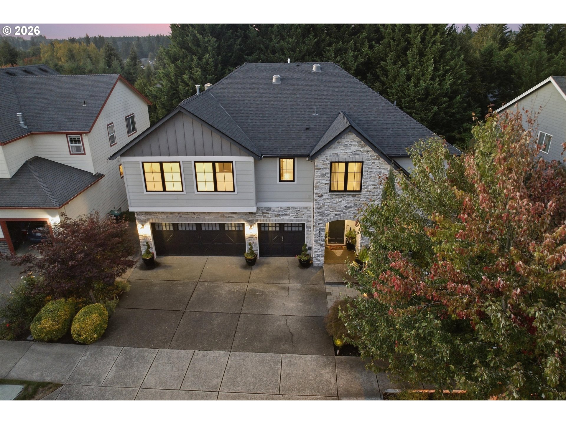 Two-story gray house with stone accents, three-car garage, and illuminated windows, viewed from above at dusk.