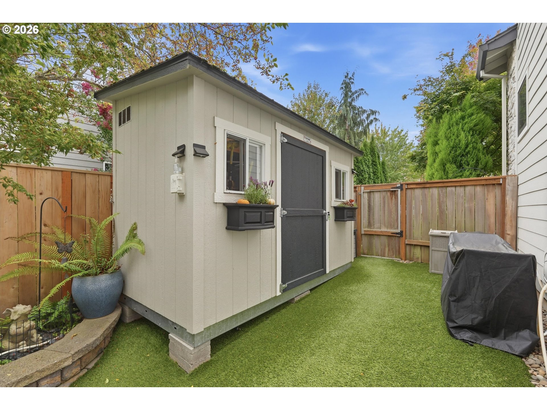 Beige backyard storage shed beside artificial turf, wooden fencing, potted ferns, and a covered grill.