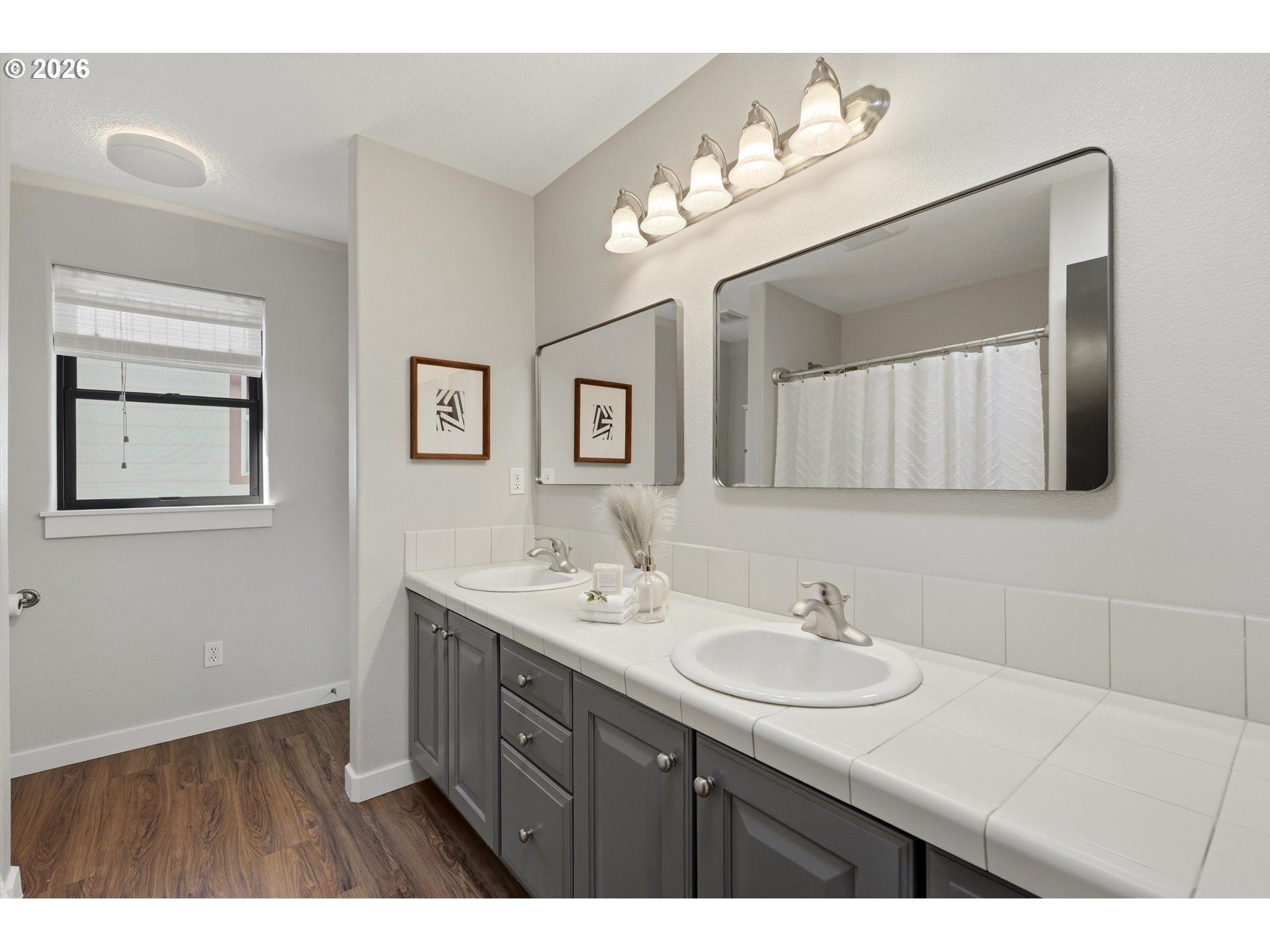 Bathroom with a double-sink vanity, gray cabinets, large mirrors, and wood-look flooring.