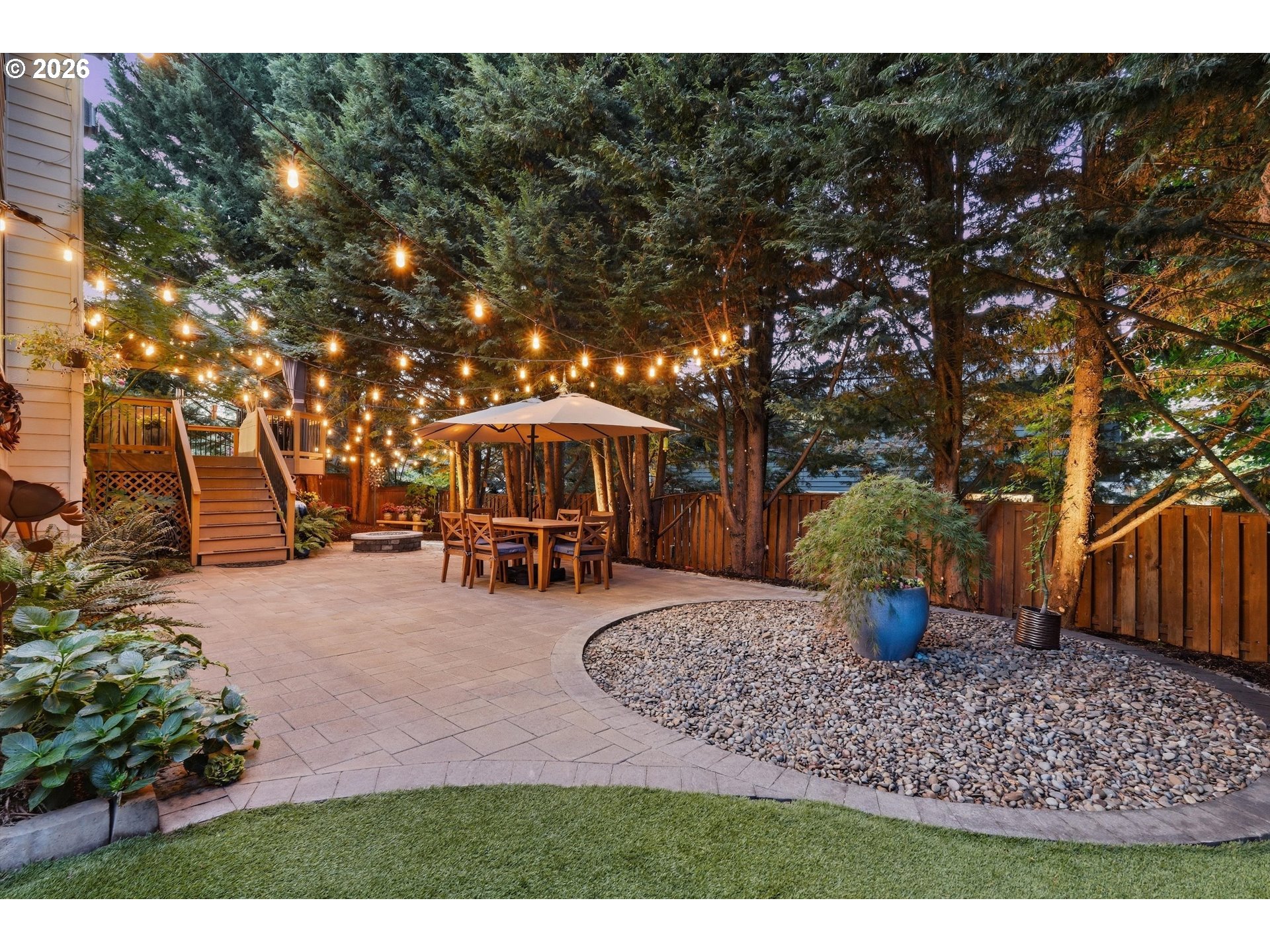 Backyard patio with string lights, outdoor dining set, raised deck, and curved pebble garden bed.