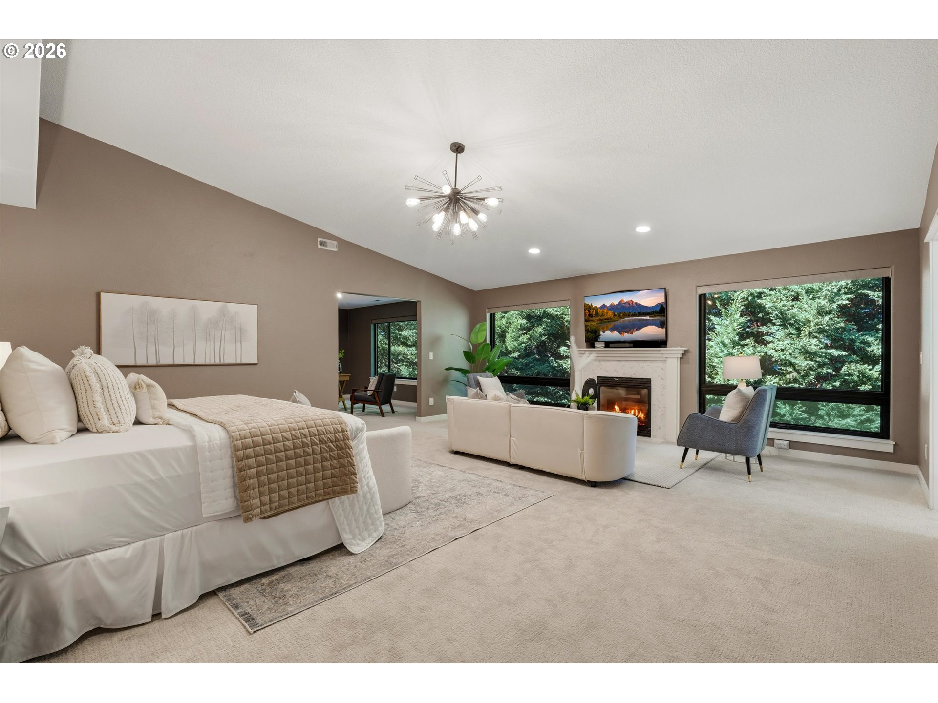 Furnished bedroom suite with a fireplace sitting area and large windows overlooking greenery.