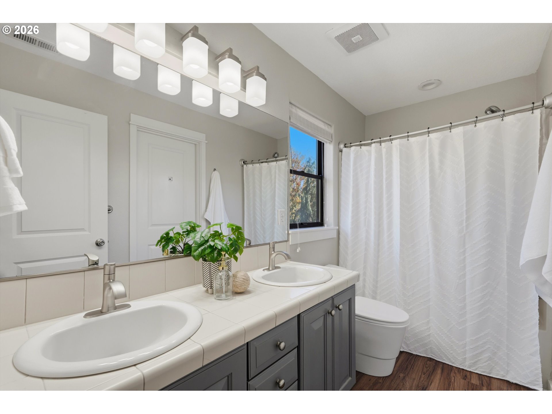 Bathroom with a tiled double-sink vanity, dark cabinets, large mirror, and white shower curtain.