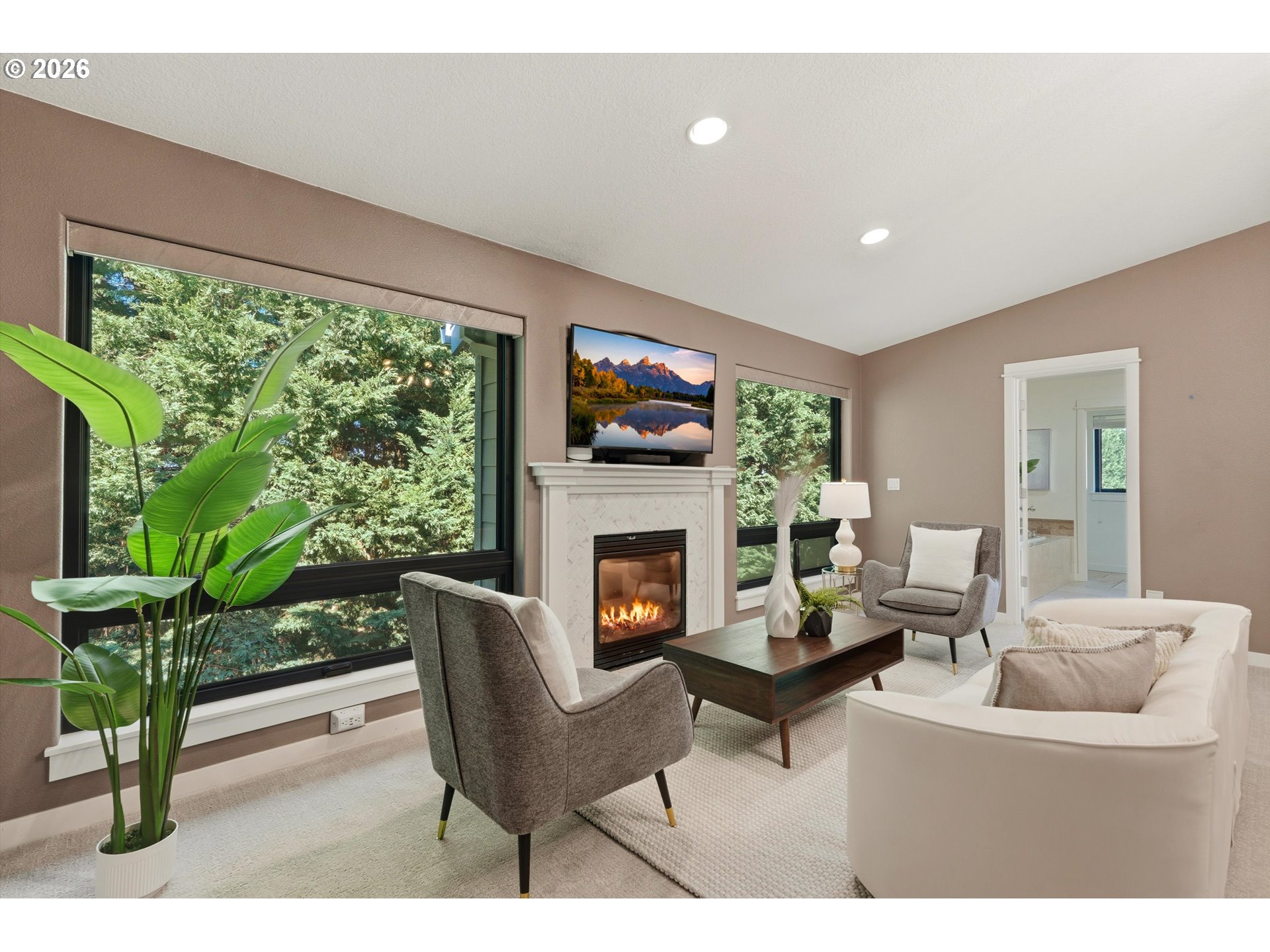 Staged living room with a fireplace, large tree-facing windows, and neutral seating.