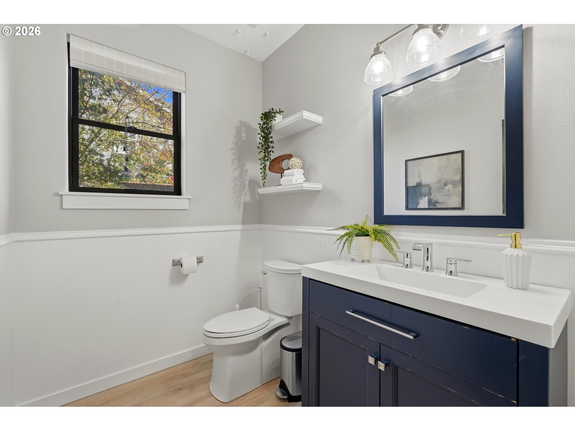 Bright bathroom with a navy vanity, white sink, large mirror, and window overlooking leafy trees.