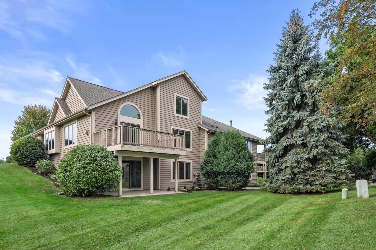 Taupe-sided home with a raised deck and arched window beside a sloping lawn and mature trees.