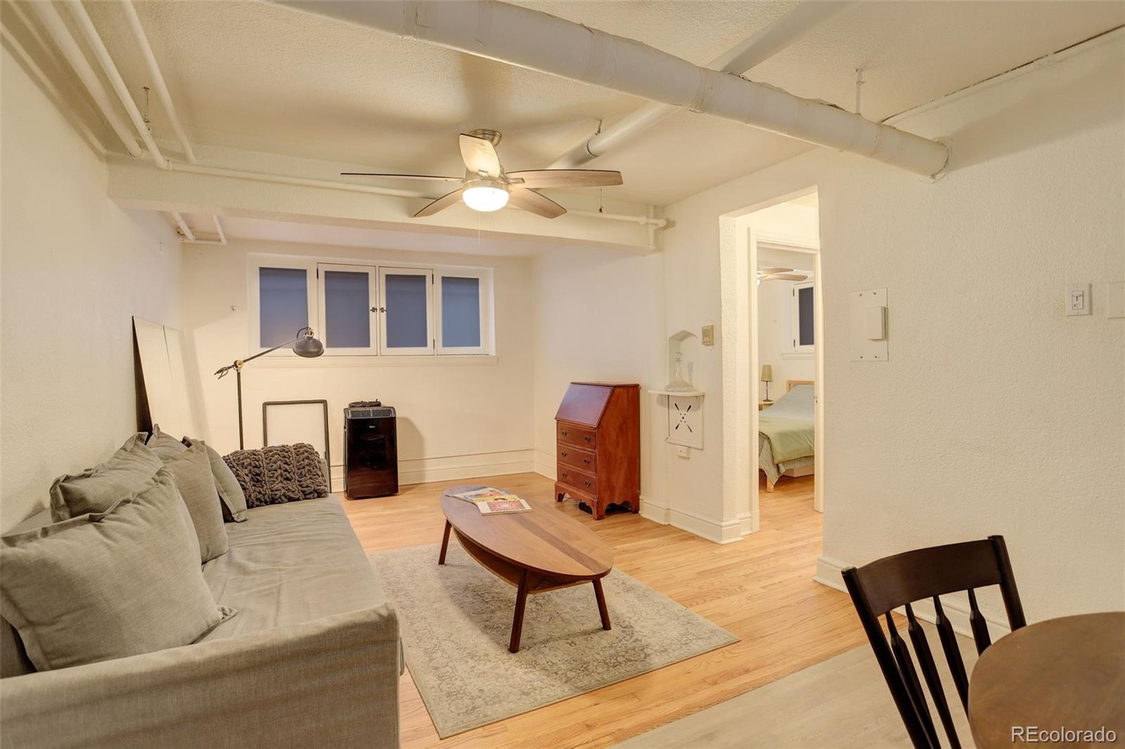 Living room with a gray sofa, hardwood floors, ceiling fan, and exposed ceiling pipes.