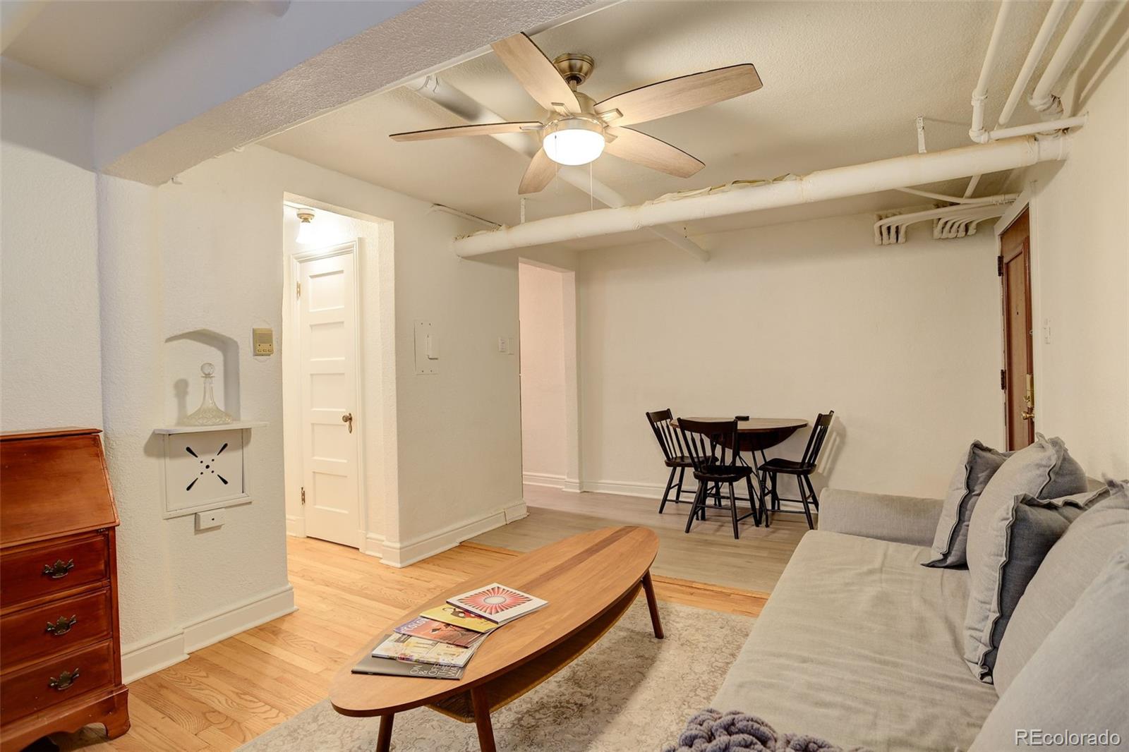 Furnished living room with a gray sofa, oval coffee table, dining nook, and exposed ceiling pipes.