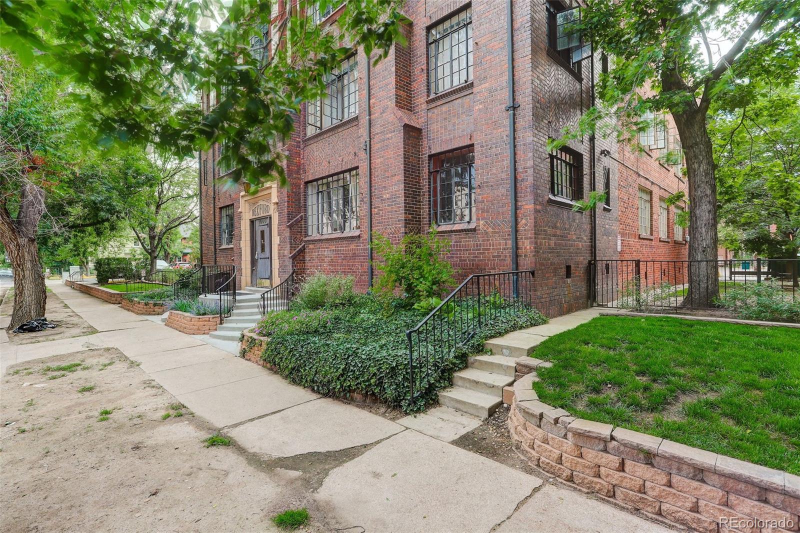 Brick apartment building with a landscaped entrance, concrete steps, and mature trees.