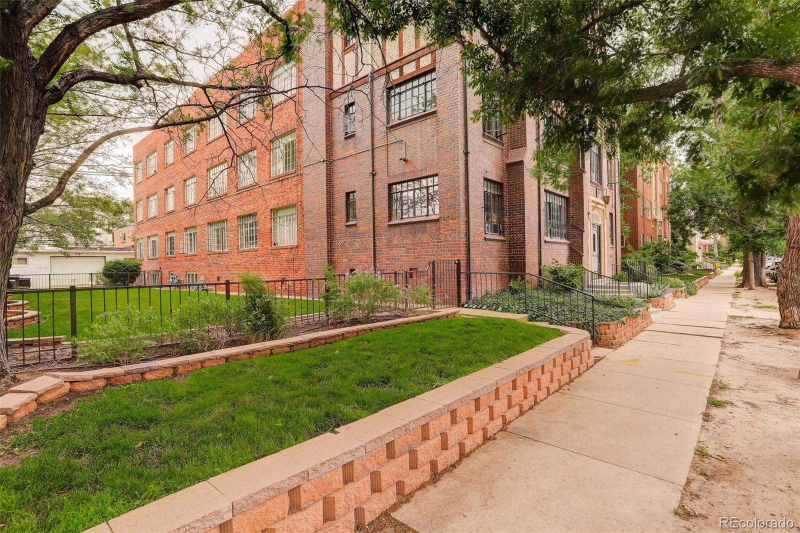 Red brick apartment building beside a fenced lawn and tree-lined sidewalk.