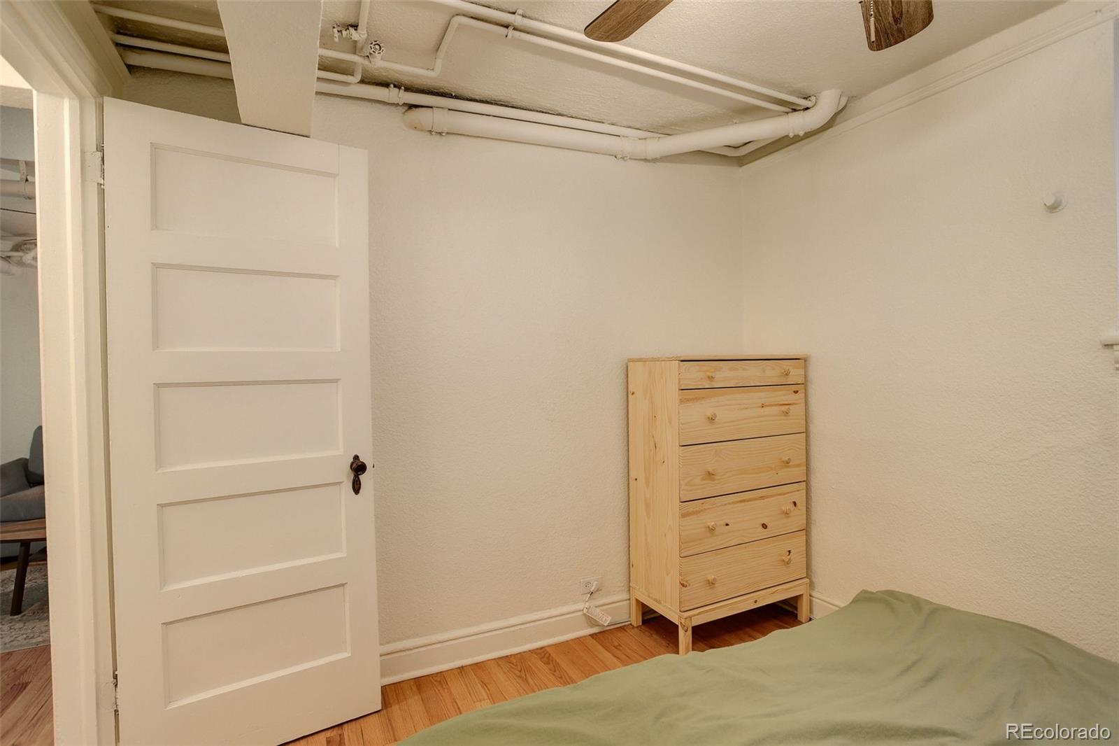 Bedroom with a green bed, light wood dresser, hardwood floor, and exposed ceiling pipes.