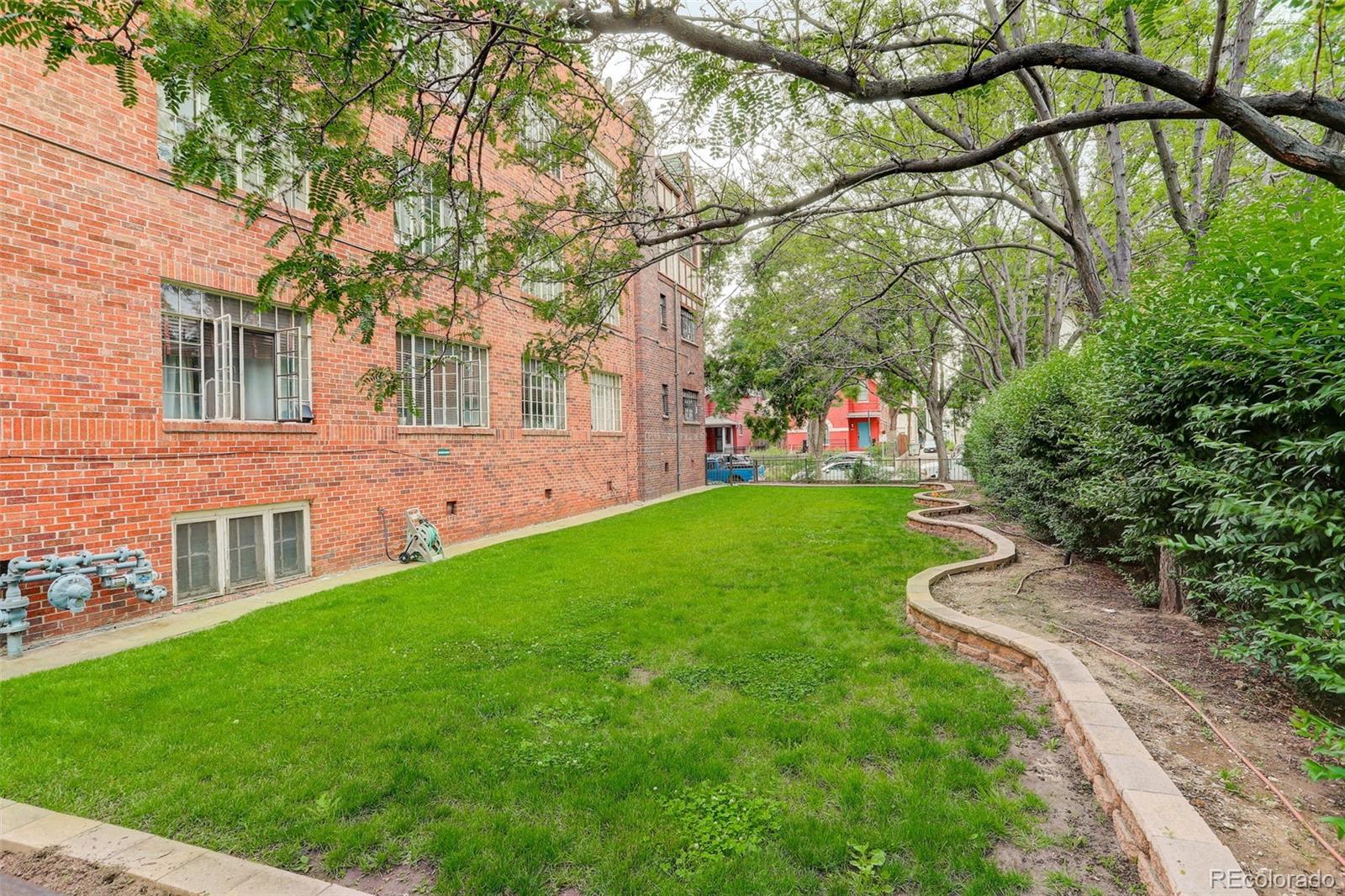 Apartment courtyard with a green lawn, mature trees, and curved garden beds beside a red-brick building.