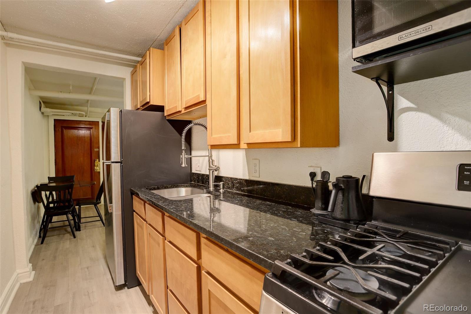 Narrow kitchen with light wood cabinets, dark countertops, stainless steel appliances, and a small dining area.