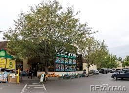 Natural Grocers storefront beneath a leafy tree, with outdoor produce displays and parked cars.