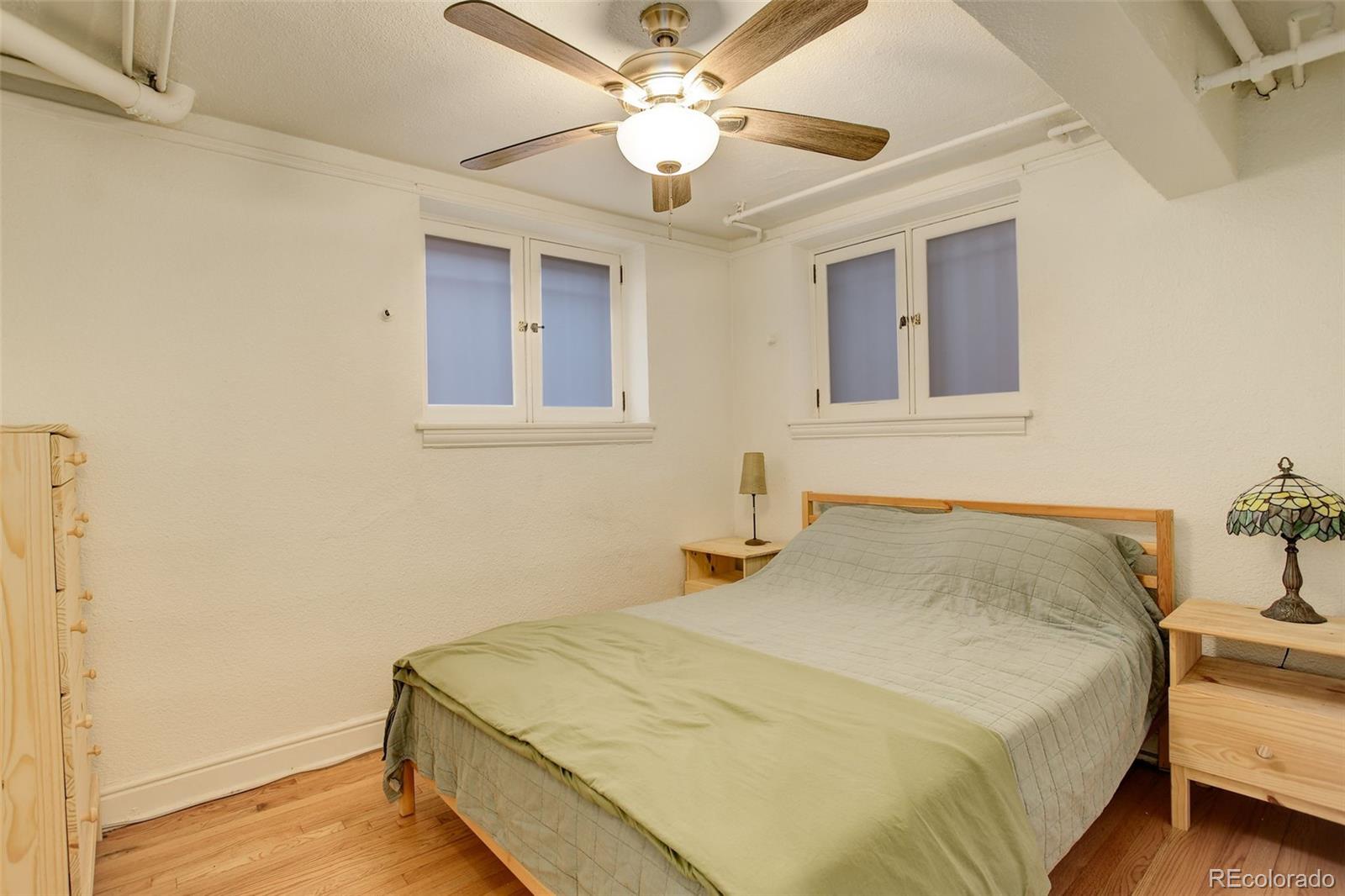 Bedroom with a wooden bed, hardwood floors, two high windows, and a ceiling fan.