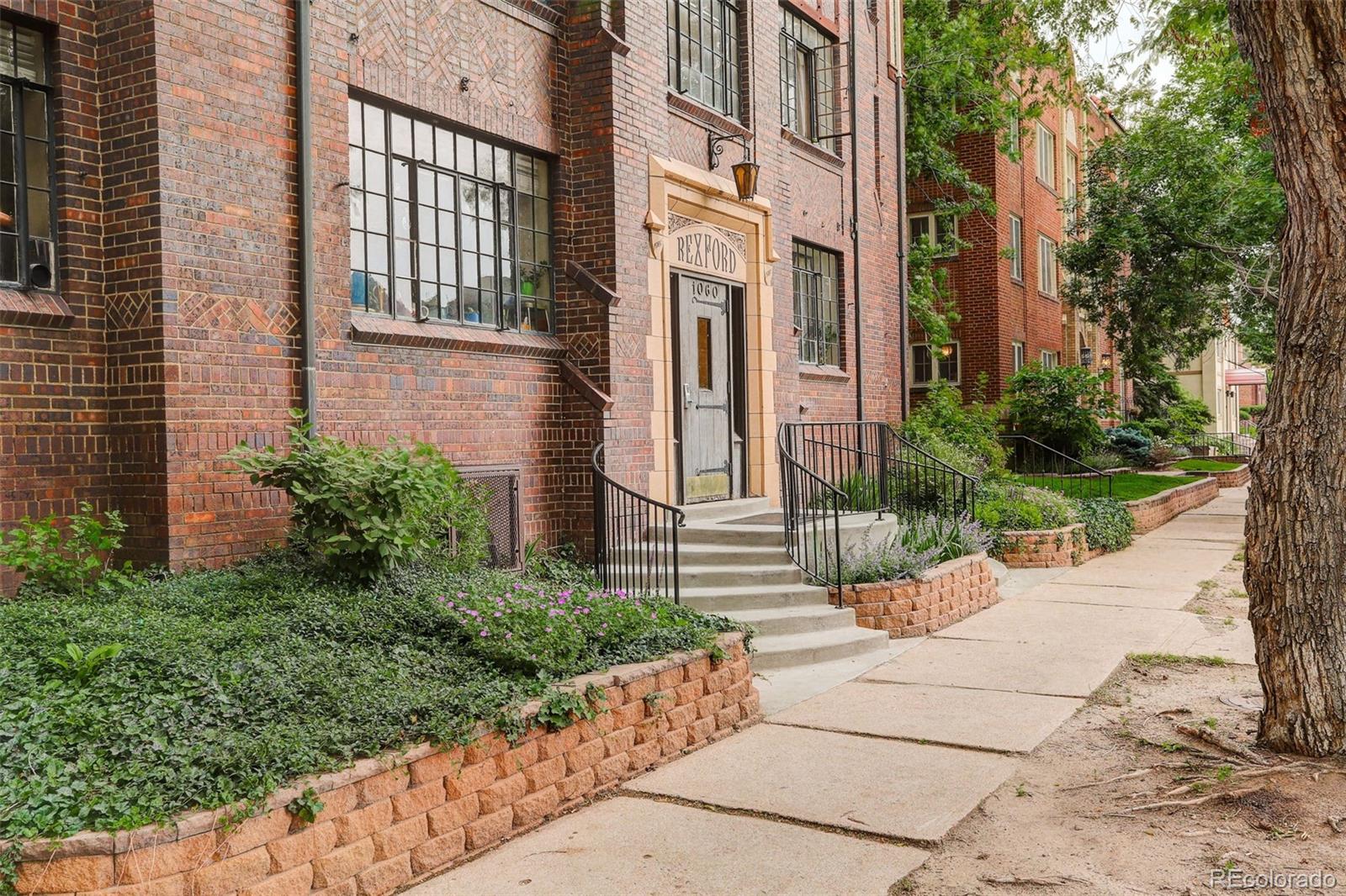 Red brick apartment building entrance with steps, black railings, and landscaped beds beside the sidewalk.
