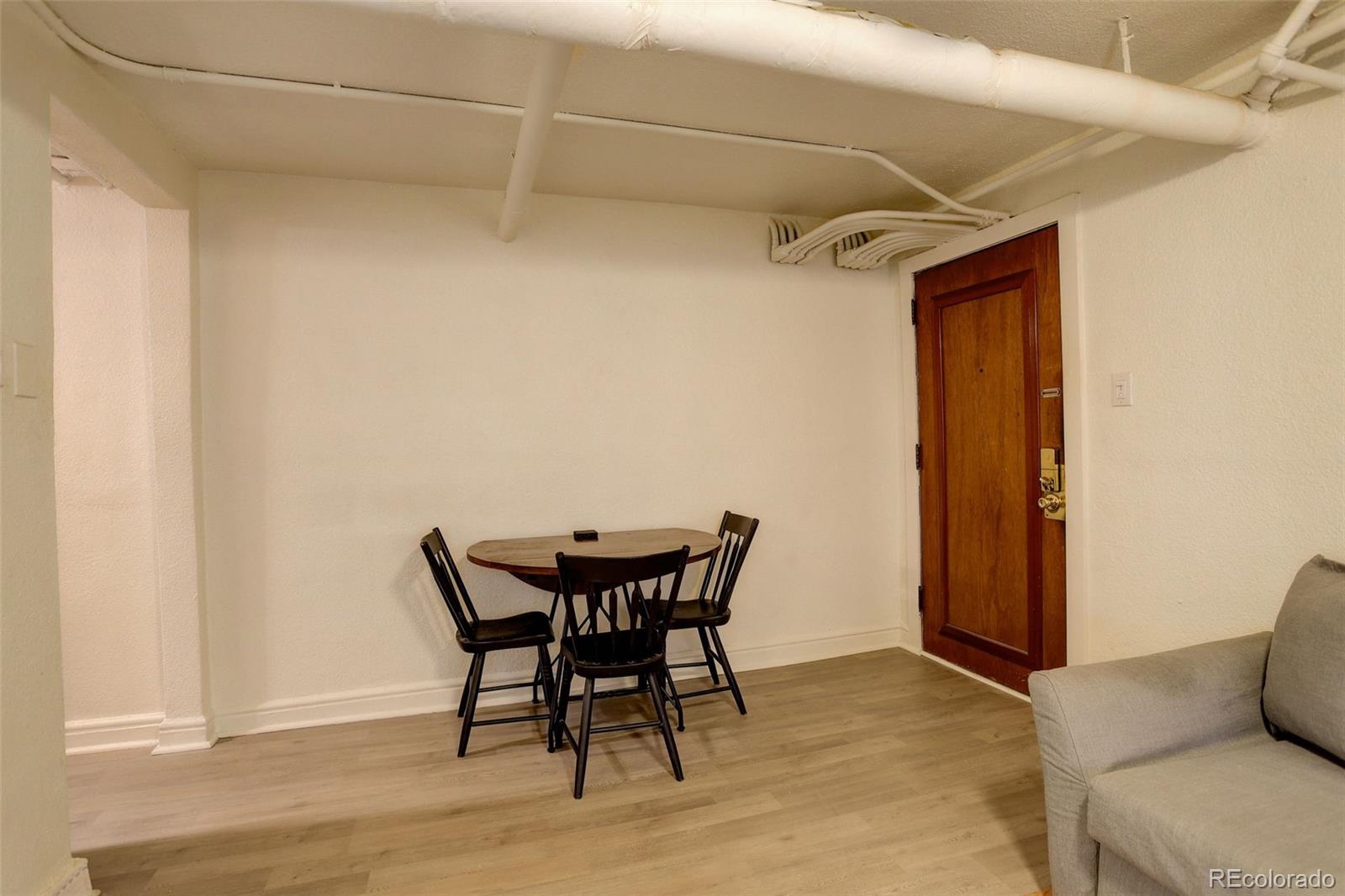 Dining area with a round table and chairs, exposed ceiling pipes, and a wooden entry door.