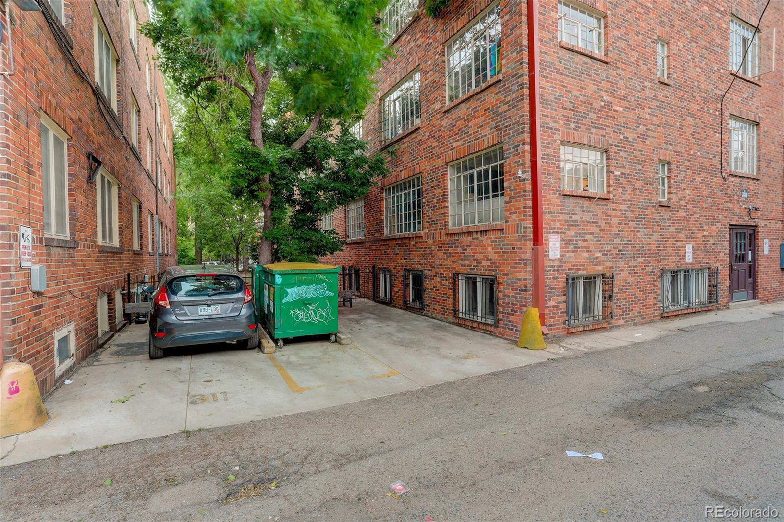 Brick apartment building beside a tree-lined alley with a parked car and green dumpster.