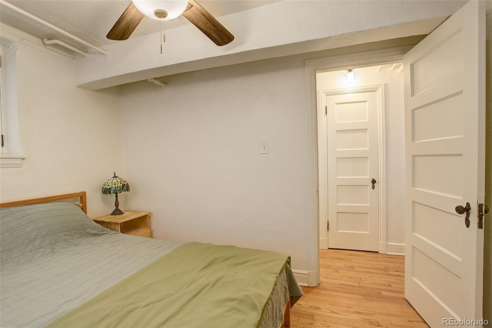 Bedroom with a ceiling fan, stained-glass bedside lamp, and open door to a hardwood-floored hall.