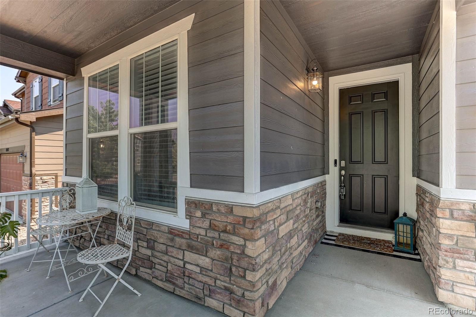 Home’s covered front porch with a black entry door, stone veneer, and white bistro set.