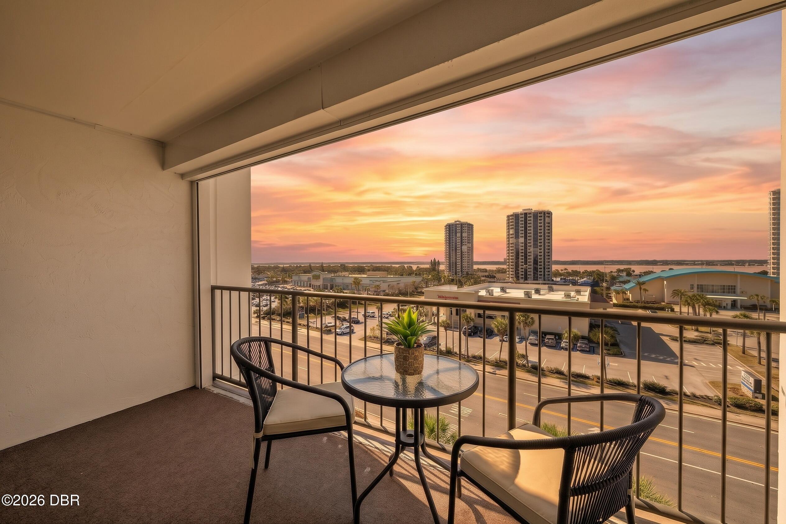 Balcony with two chairs and a table overlooking towers and water at sunset.