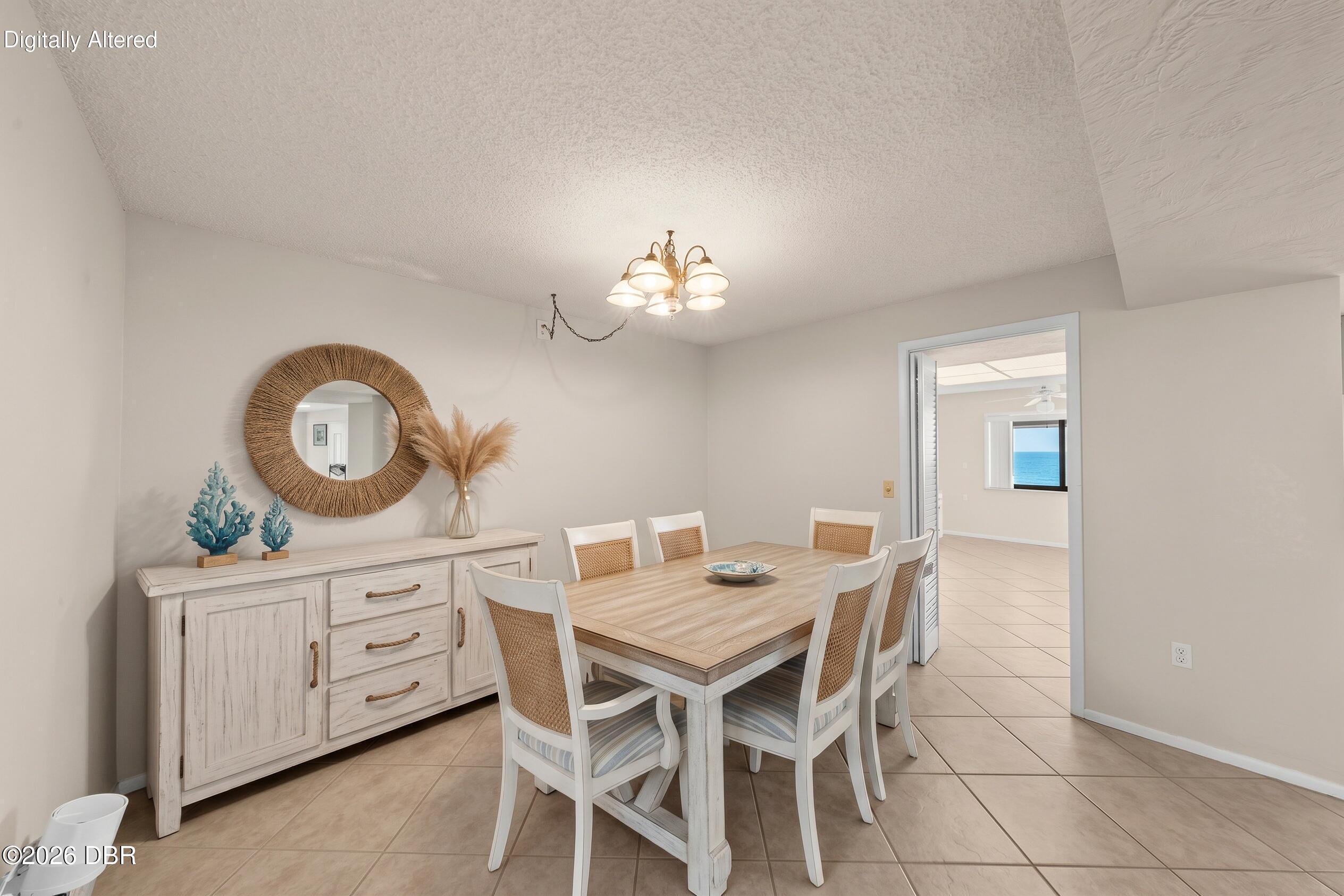 Dining room with a light wood table and woven chairs, opening to a room with an ocean view.