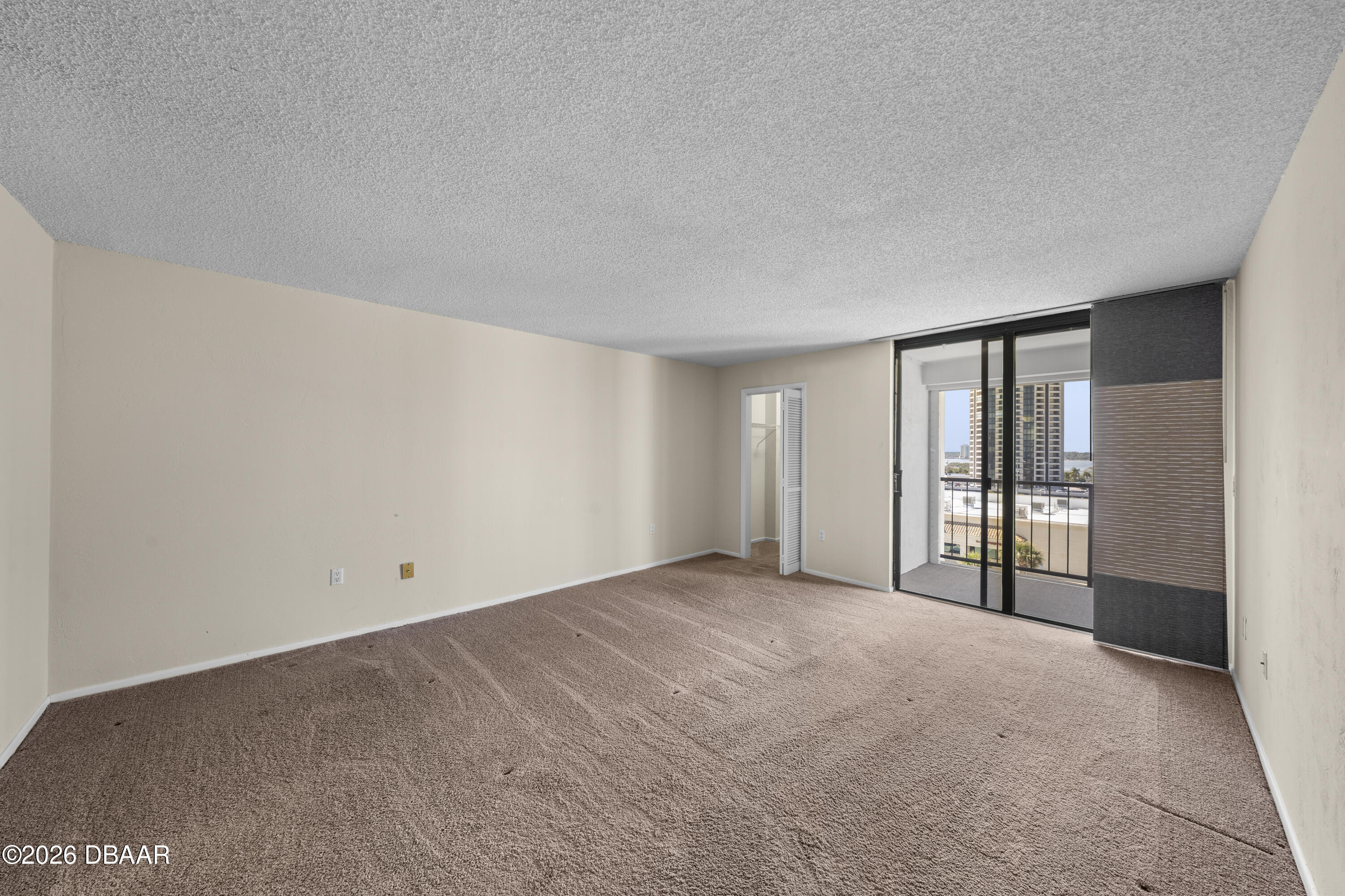 Empty carpeted bedroom with sliding glass doors opening to a balcony and views of nearby buildings.