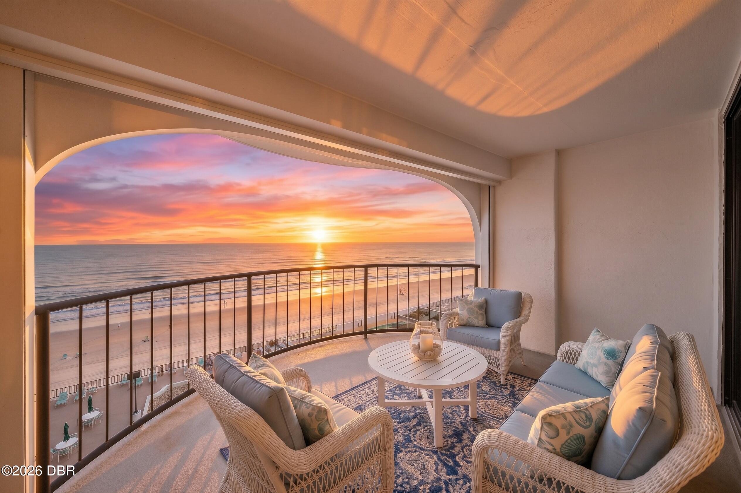 Furnished oceanfront balcony with wicker seating overlooking the beach at sunset.