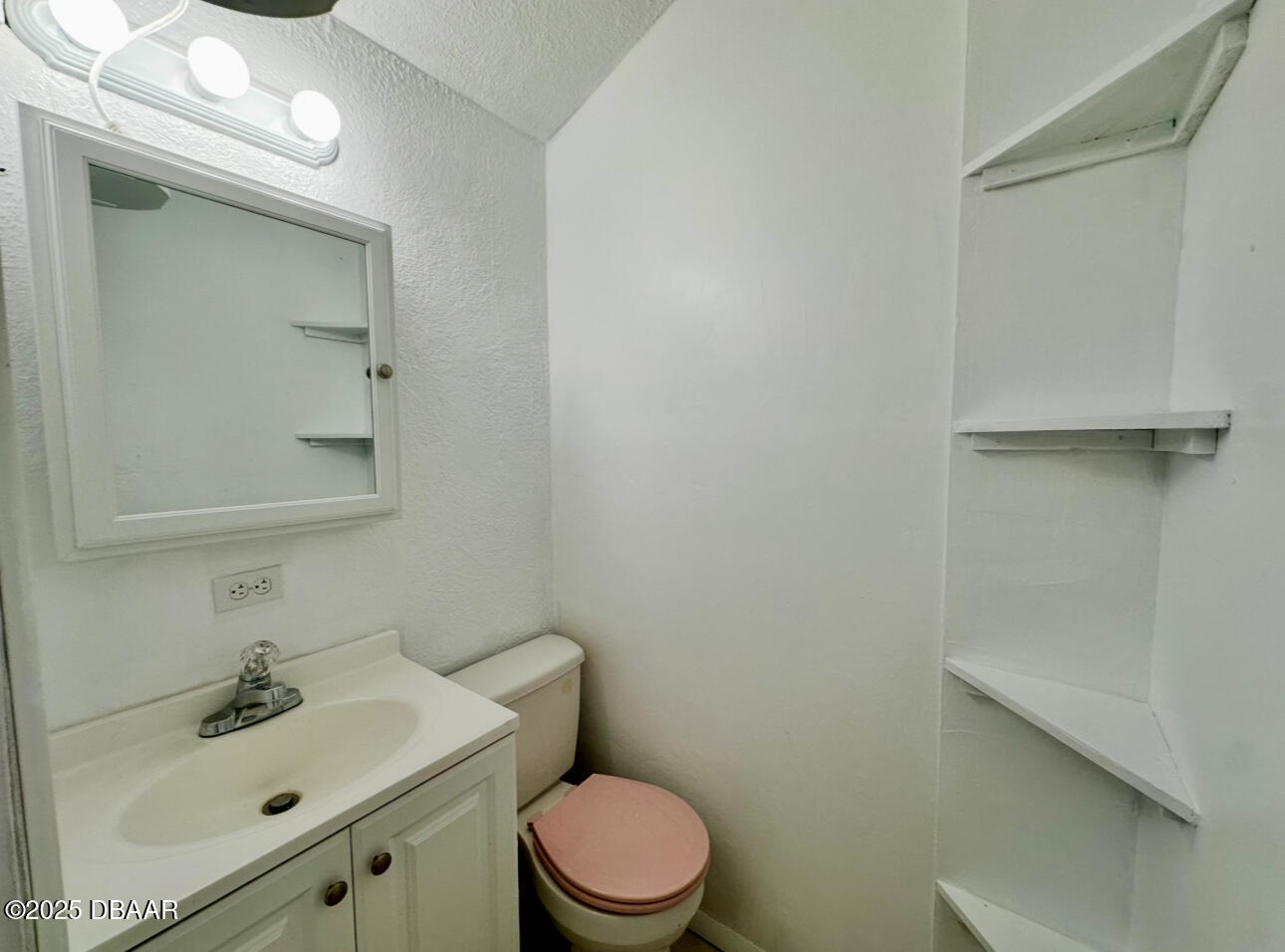 Small bathroom with a white vanity, pink toilet seat, and open corner shelves.
