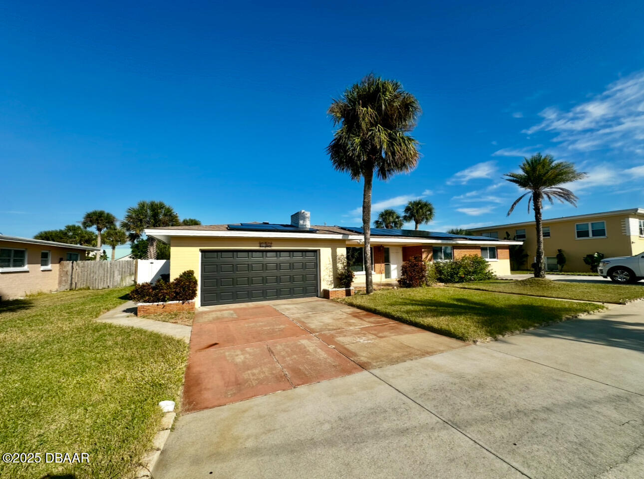 Yellow cottage with rooftop solar panels, a wide driveway, and palm trees in the yard.
