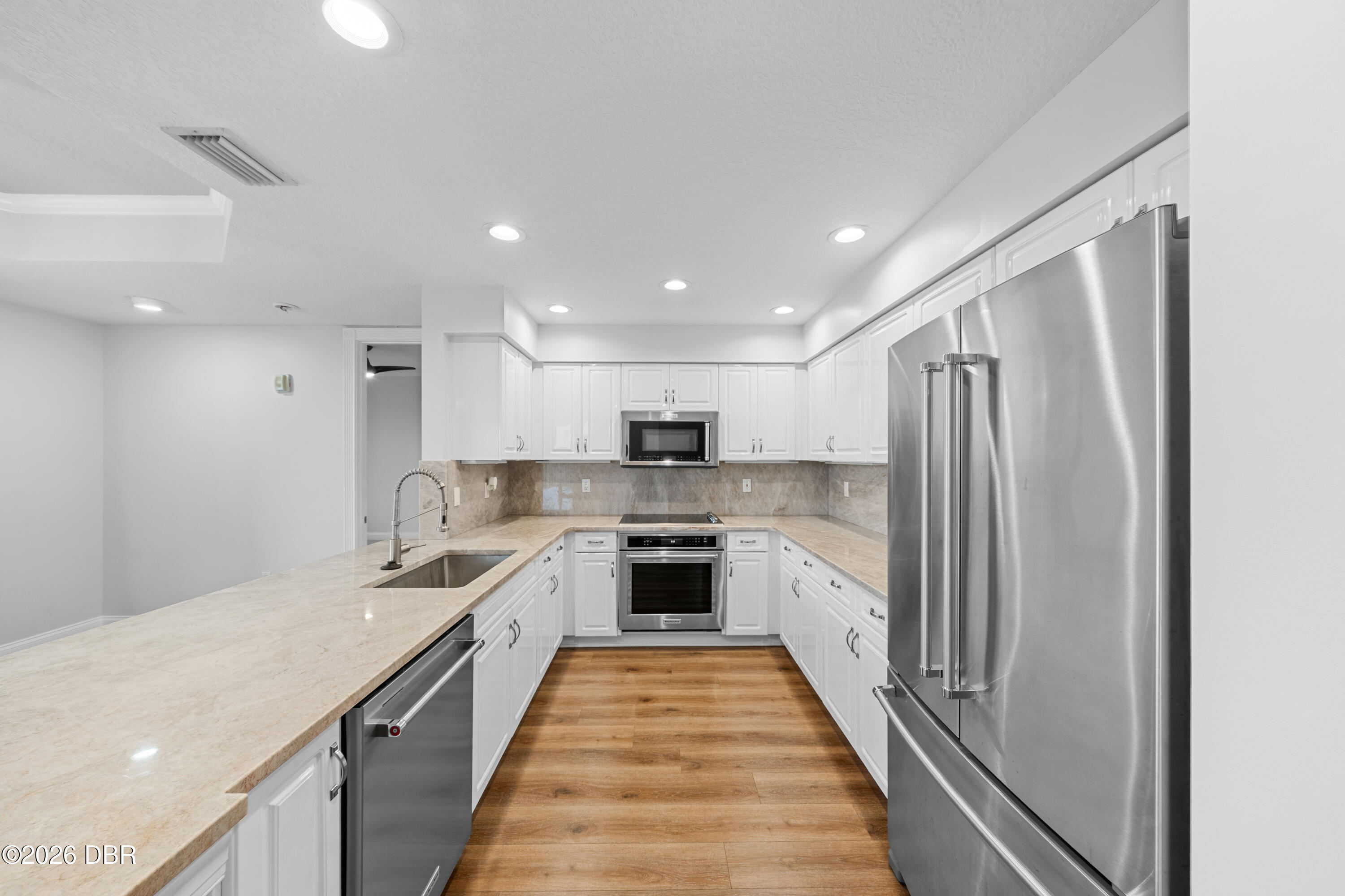 Kitchen with white cabinetry, beige stone counters, stainless appliances, and warm wood-look flooring.