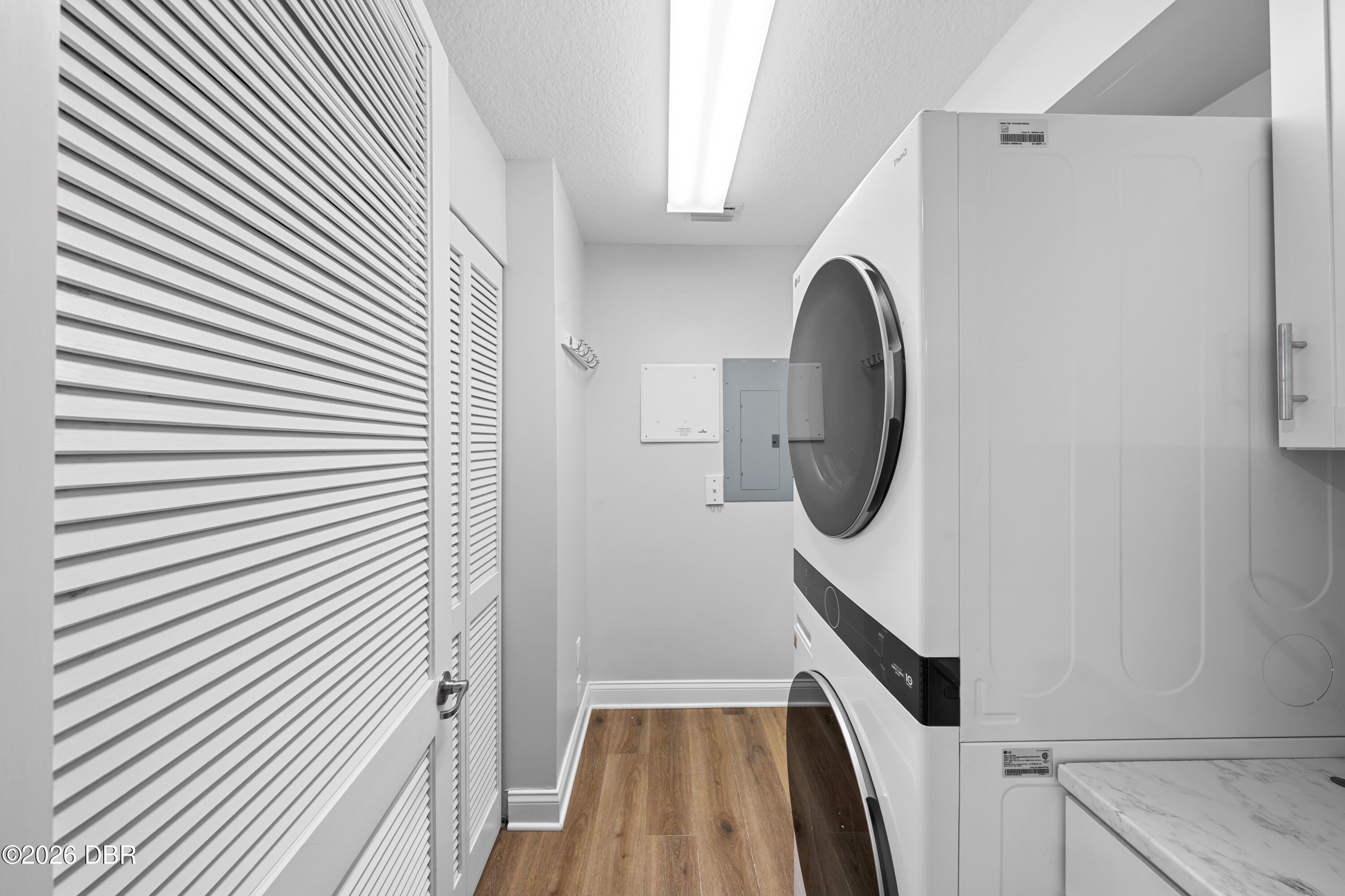 Laundry room with a stacked washer and dryer, louvered doors, and wood-look flooring.
