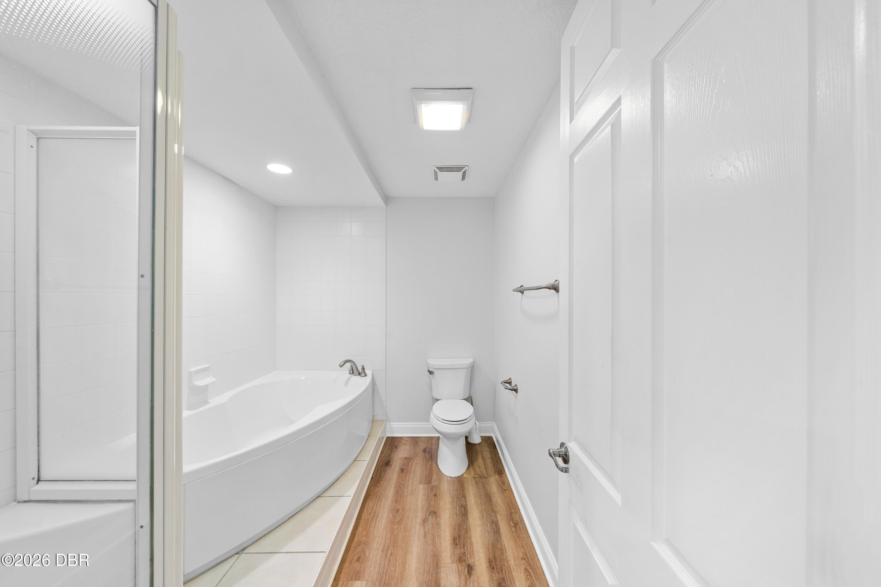 Bathroom with a white corner tub, glass shower enclosure, toilet, and wood-look flooring.