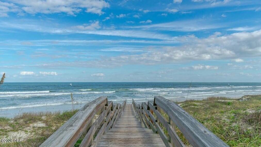 Beach boardwalk leading through coastal grass to the ocean beneath a bright, cloud-streaked sky.