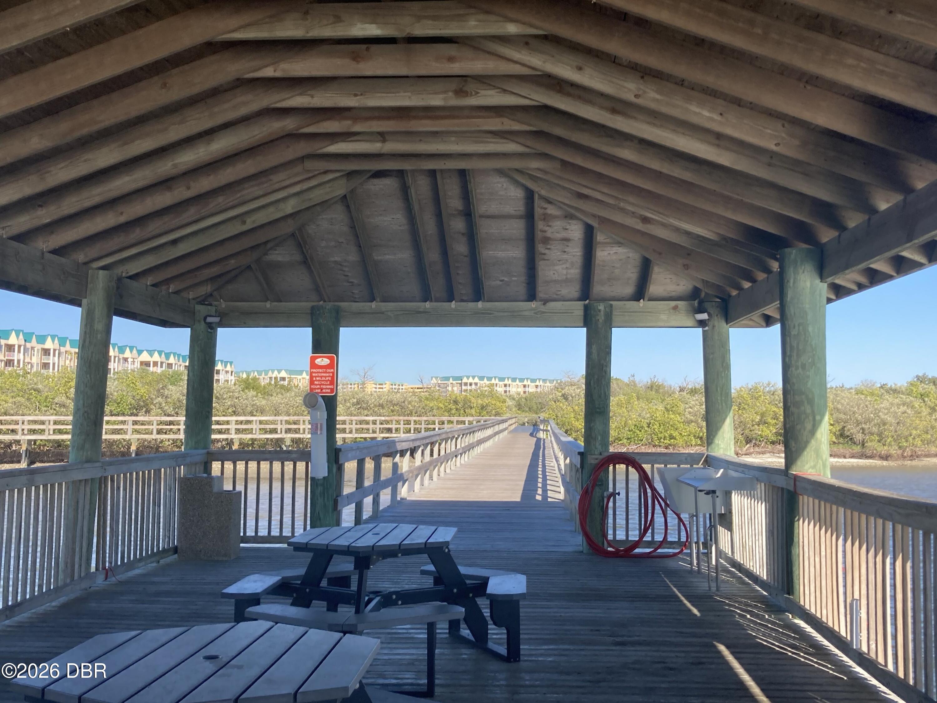 Covered wooden pier extends over the water toward a boardwalk, with picnic tables and mangroves.