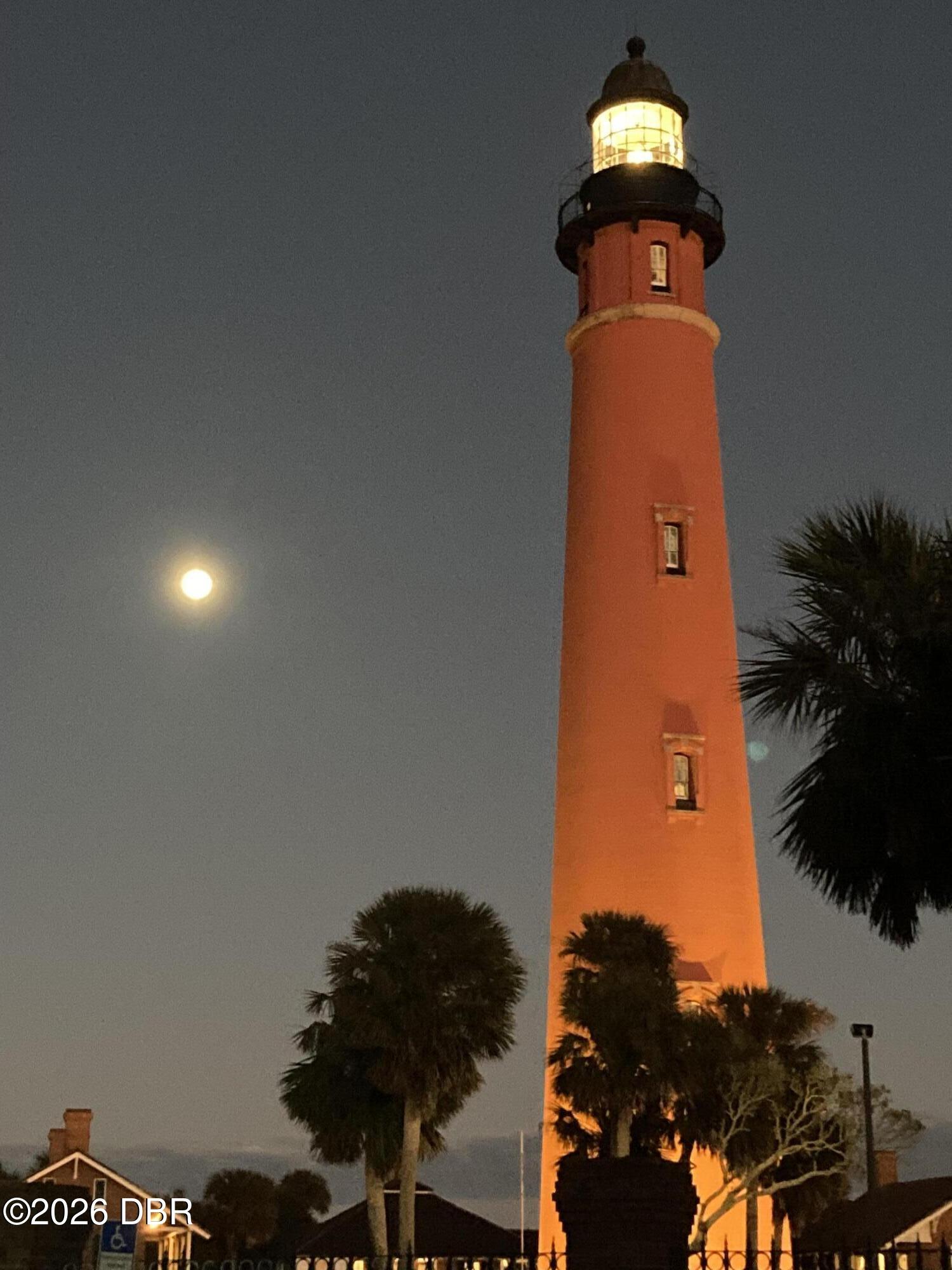 Red lighthouse illuminated beneath a full moon, framed by palm trees.