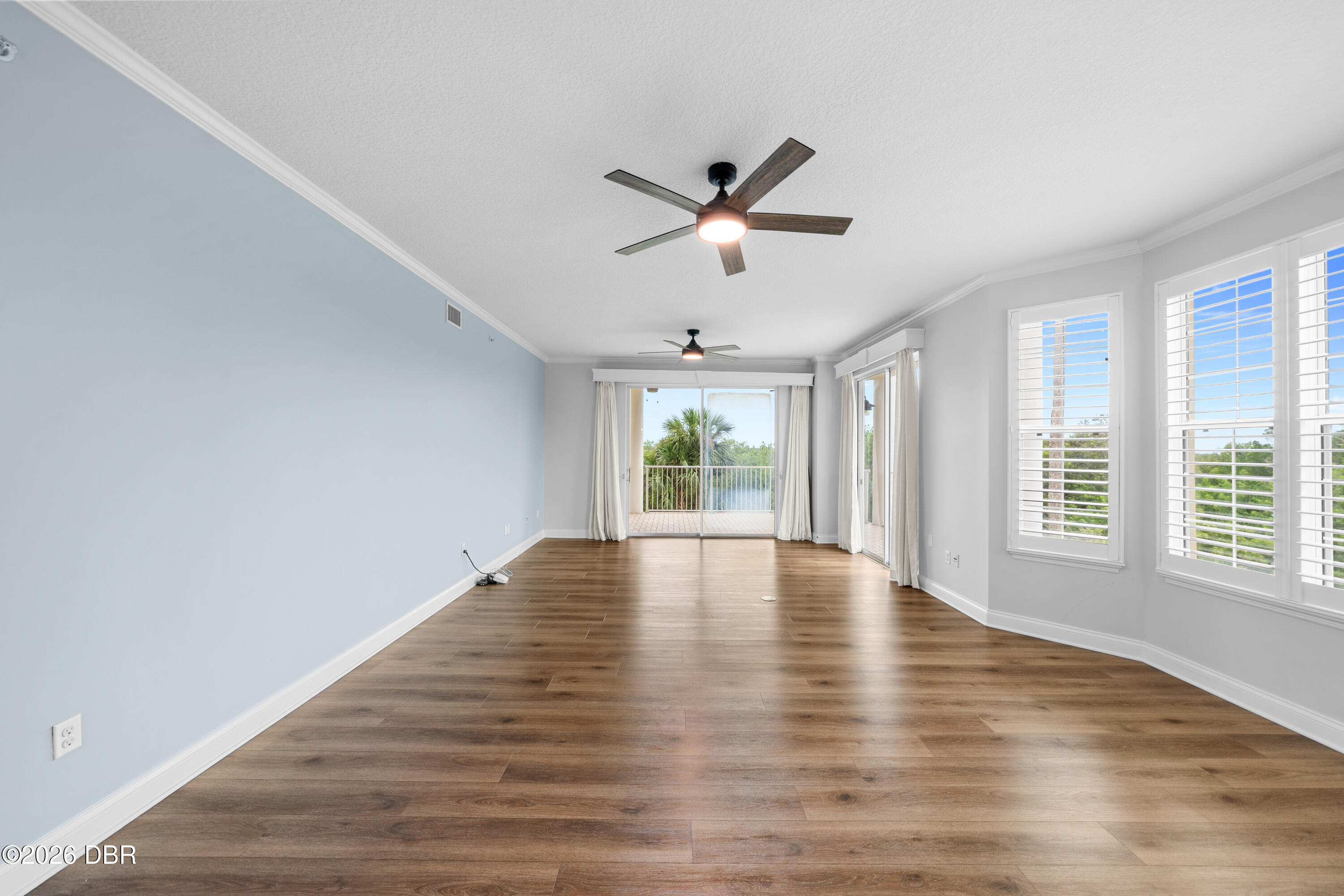 Empty living room with wood floors, ceiling fans, plantation shutters, and sliding glass doors to a balcony.
