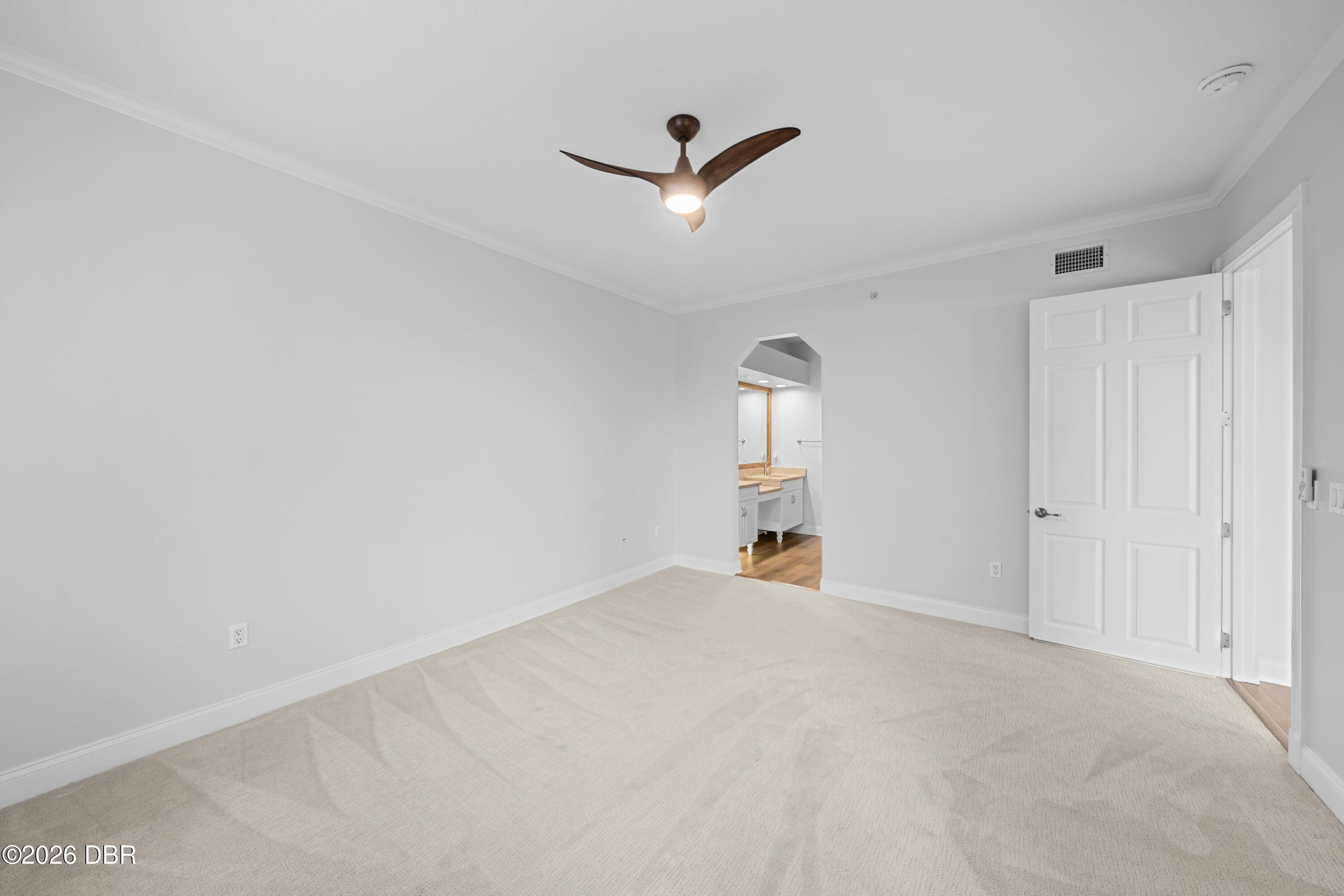 Empty bedroom with light carpet and a ceiling fan, opening to a bathroom vanity.