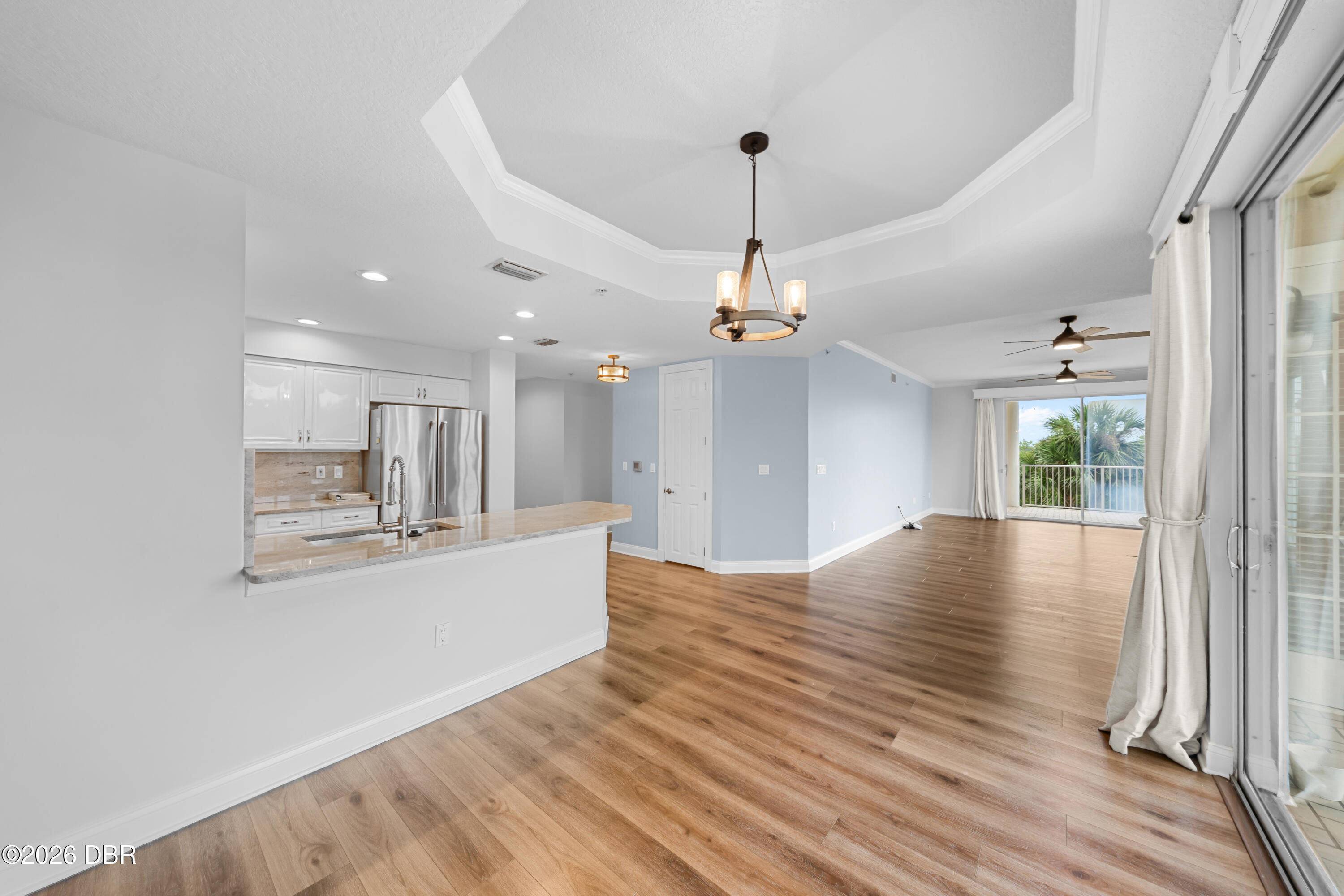 Open condo interior with kitchen peninsula, wood-look floors, tray ceiling, and sliding doors to a palm-lined balcony.